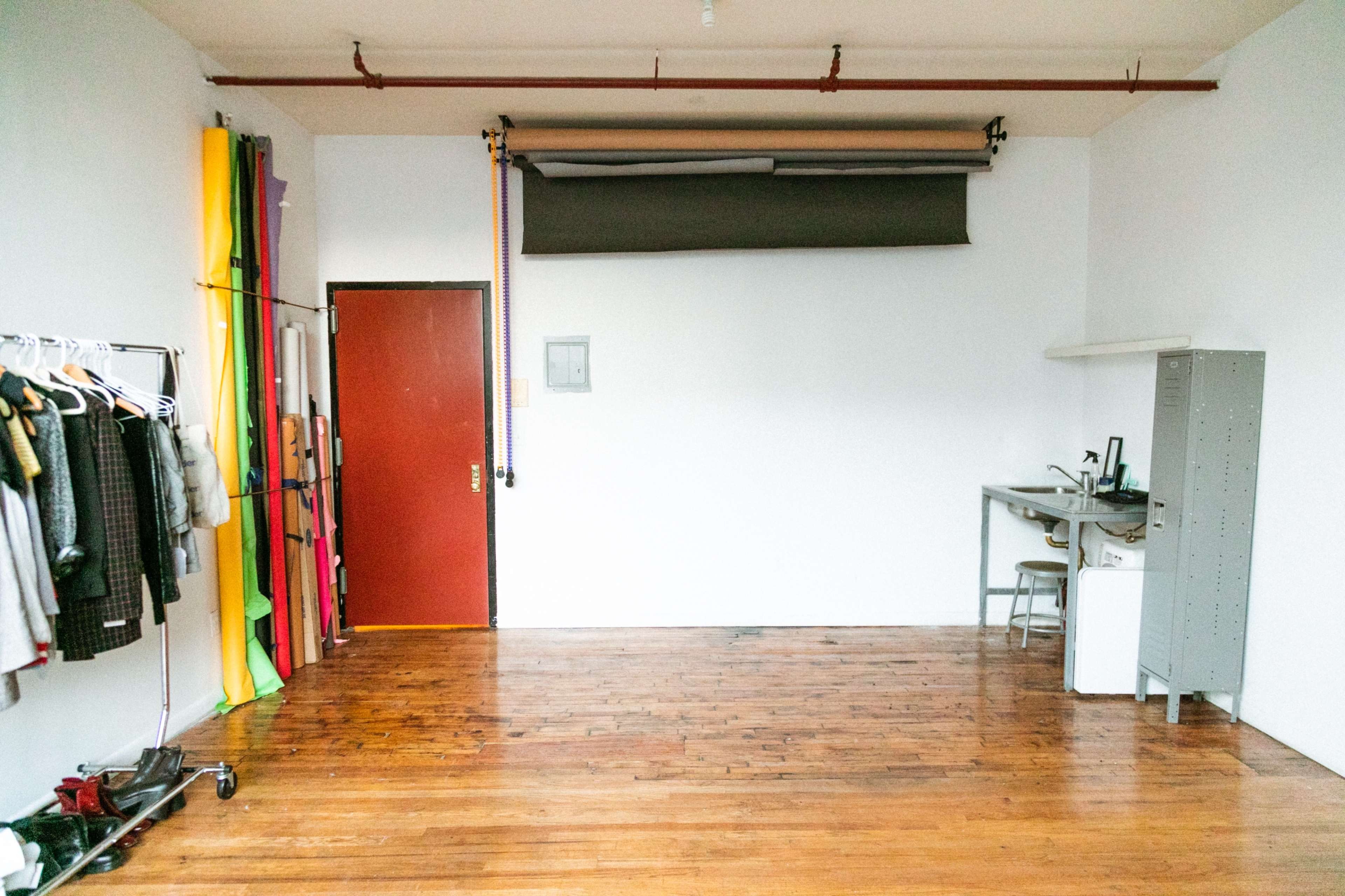 The image shows a spacious, minimally furnished studio with wooden floors, a red door, and a clothing rack filled with various garments.