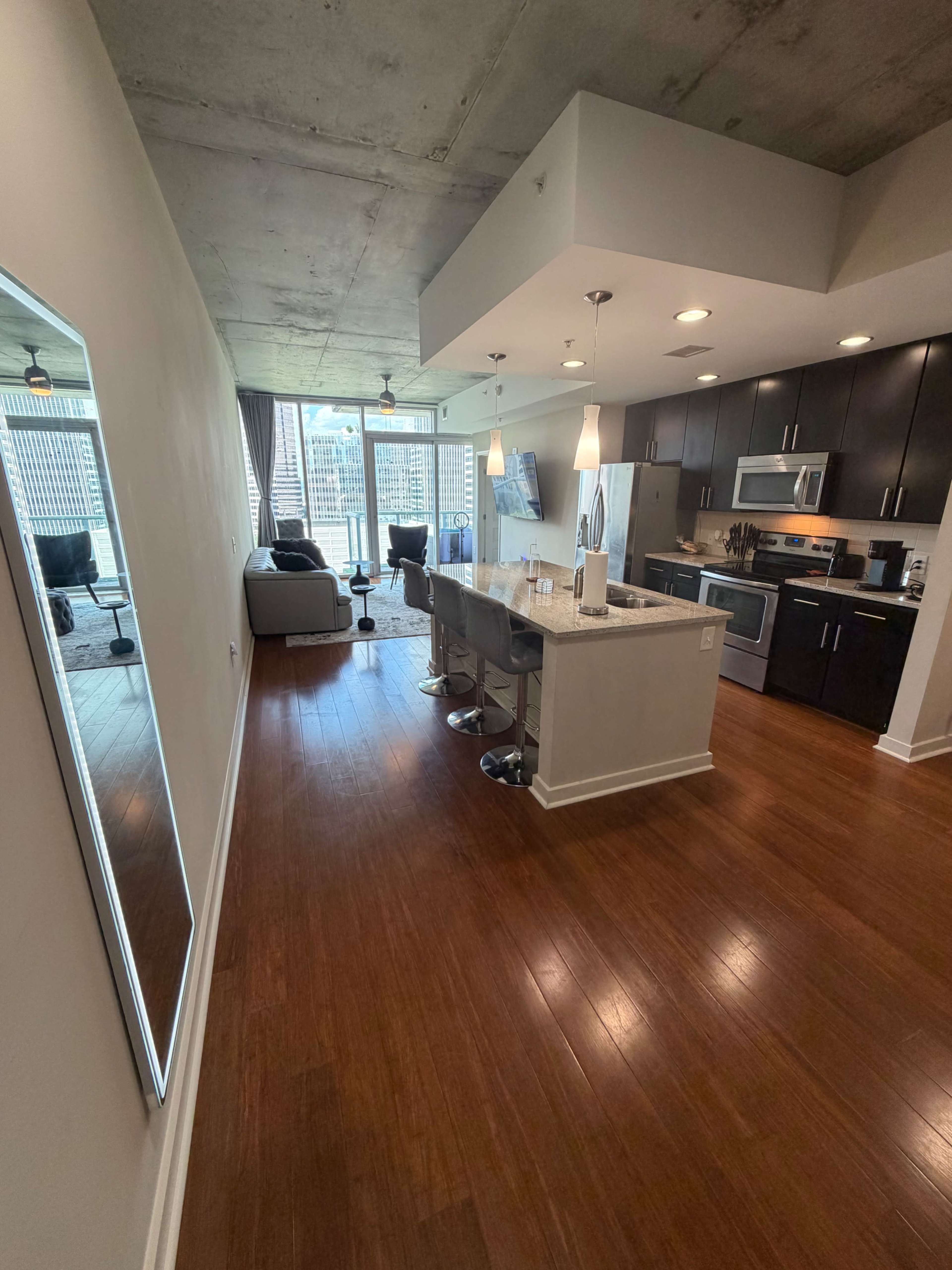 The image shows a modern, open-concept apartment featuring a kitchen with bar stools, a living area with a sofa, and large windows letting in natural light.