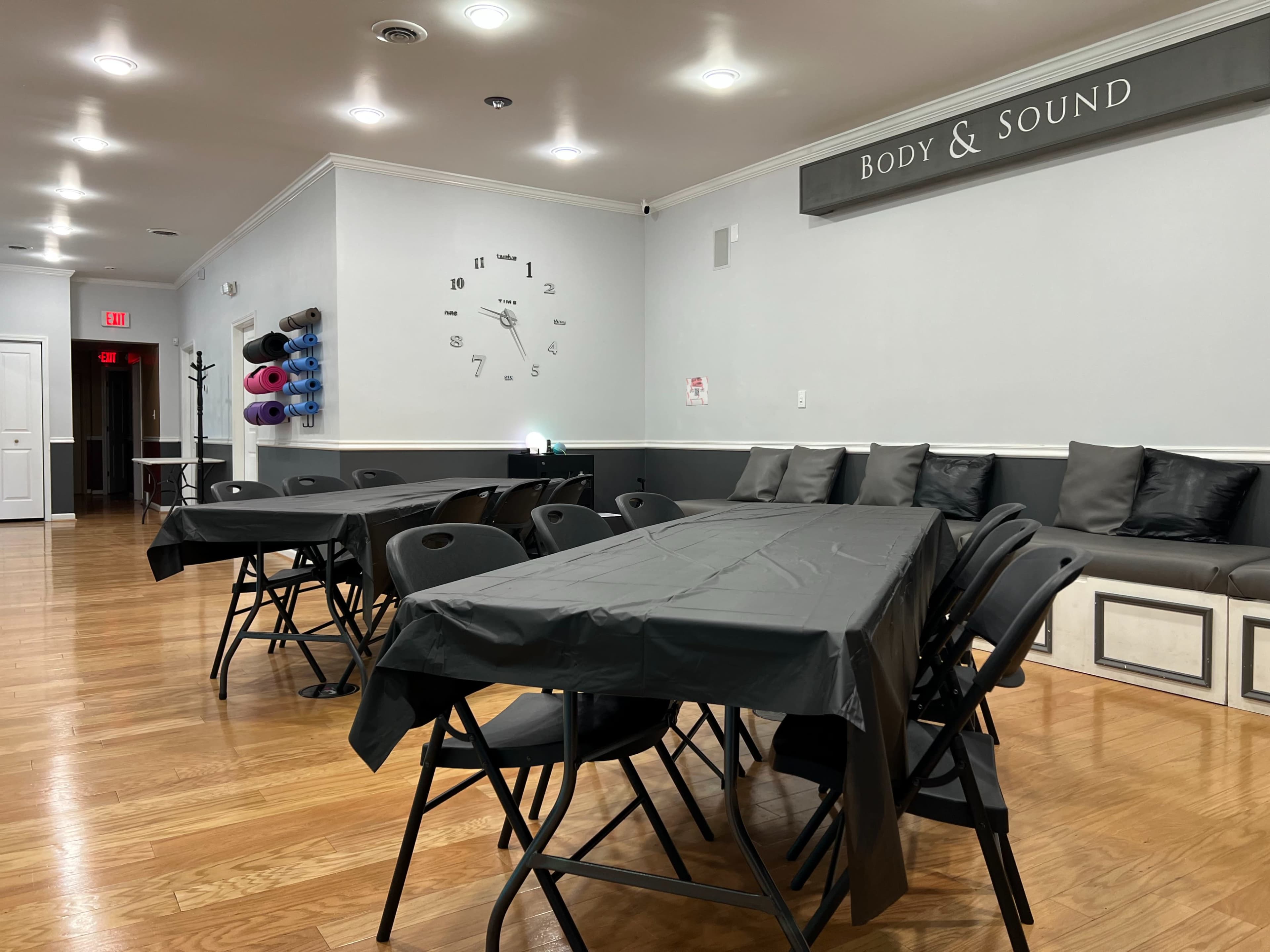 A spacious room with several black folding tables arranged for an event, a wall clock, and a seating area with gray cushions.