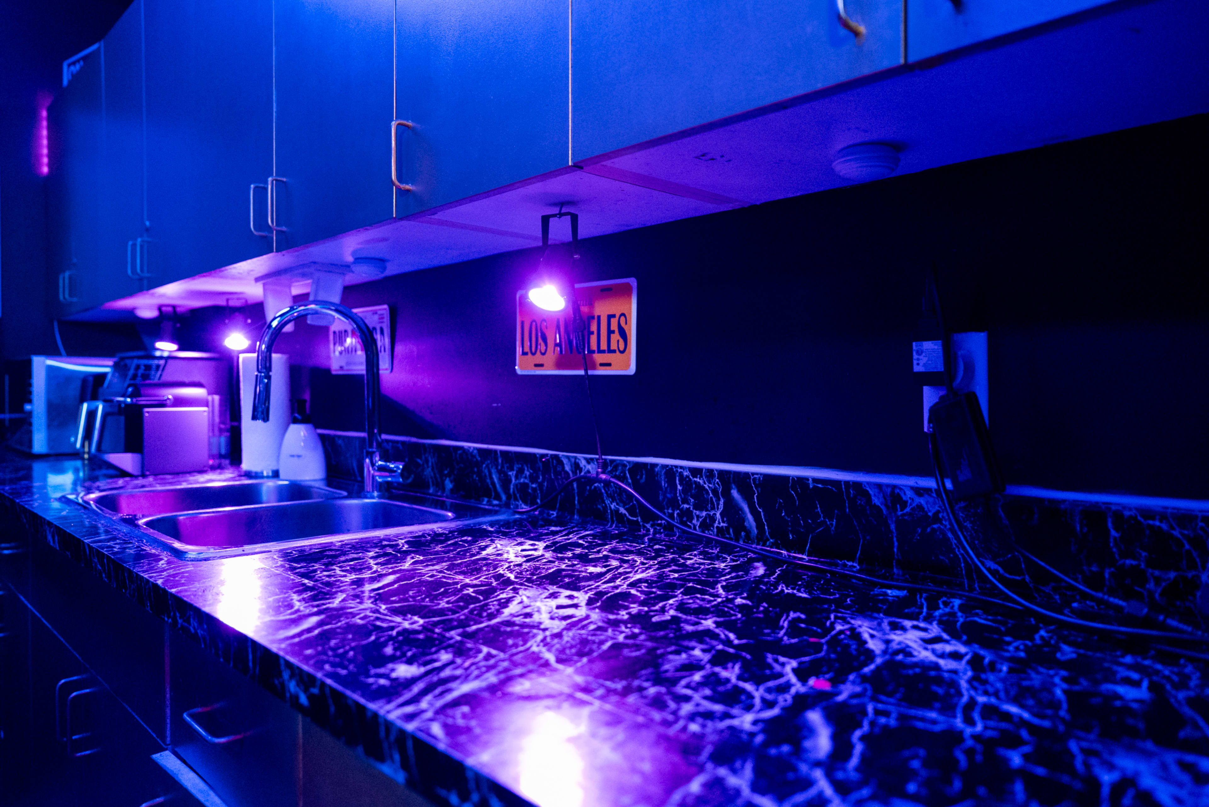 The image shows a kitchen with a dark marble countertop, illuminated by purple lights under the cabinets, and featuring a sink and various appliances.