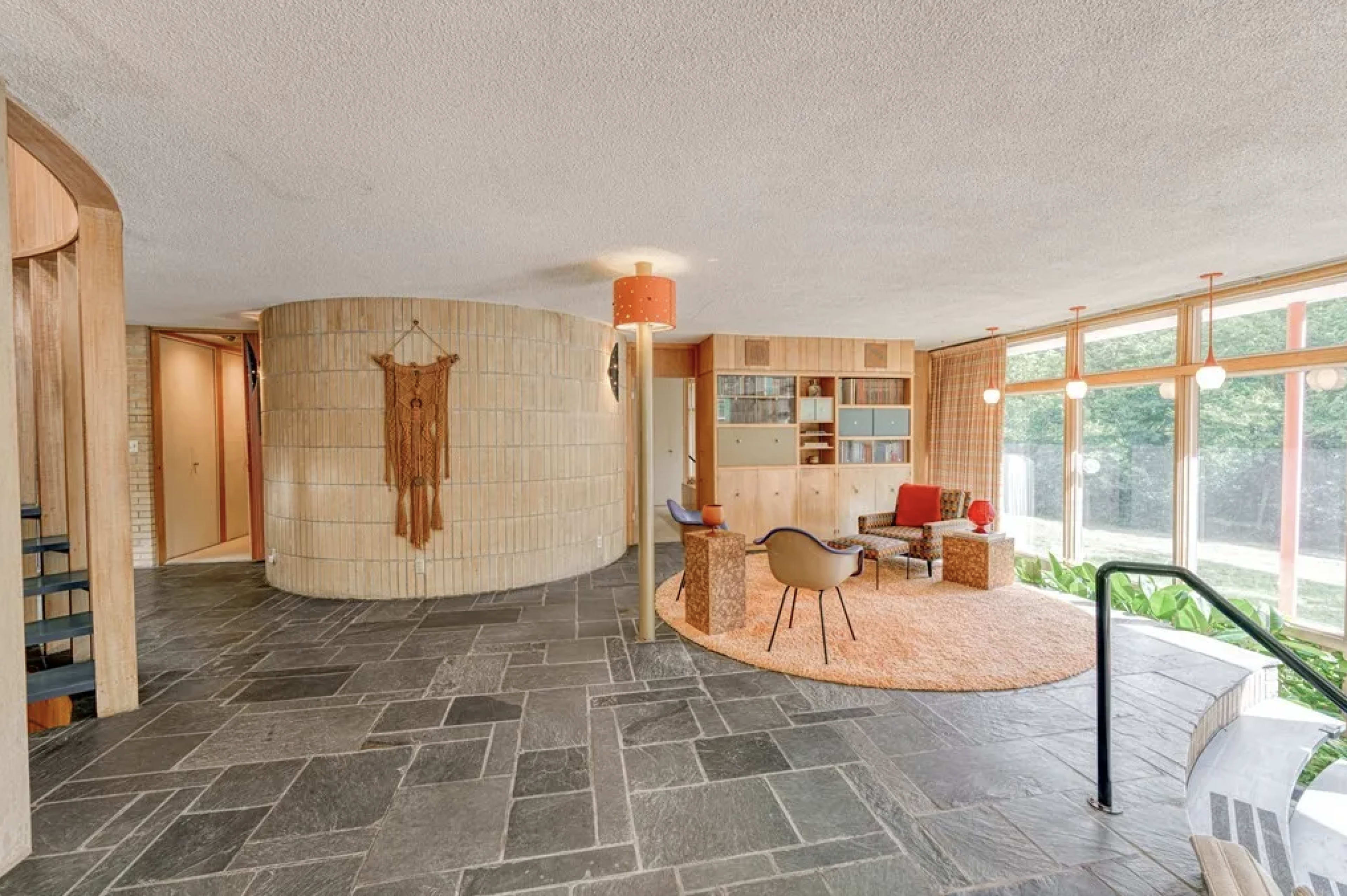 The image shows a modern interior space featuring a circular structure with stone flooring, wooden accents, and a seating area with colorful furniture and large windows.