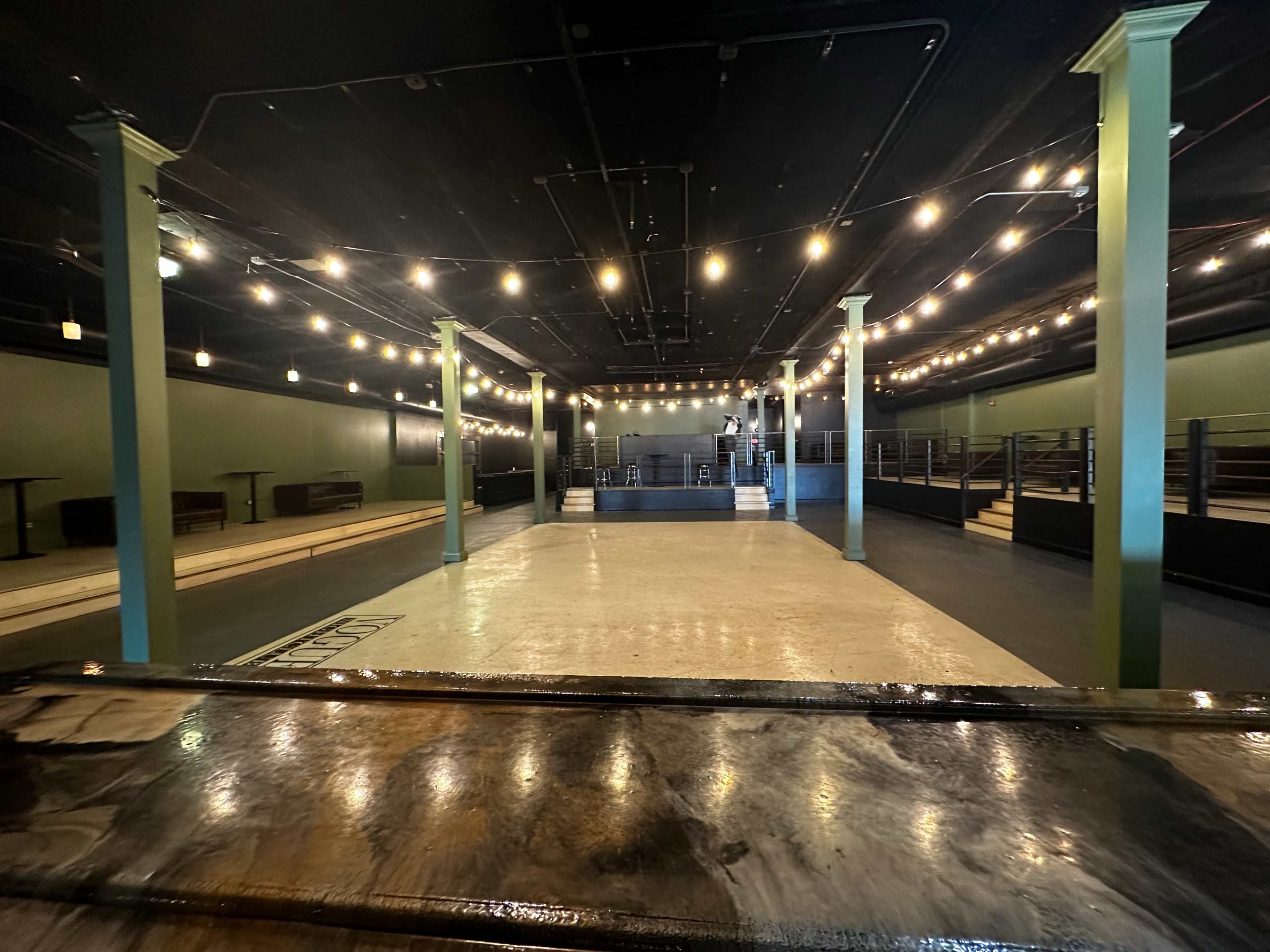 An empty event space with a polished floor, green columns, and string lights overhead.