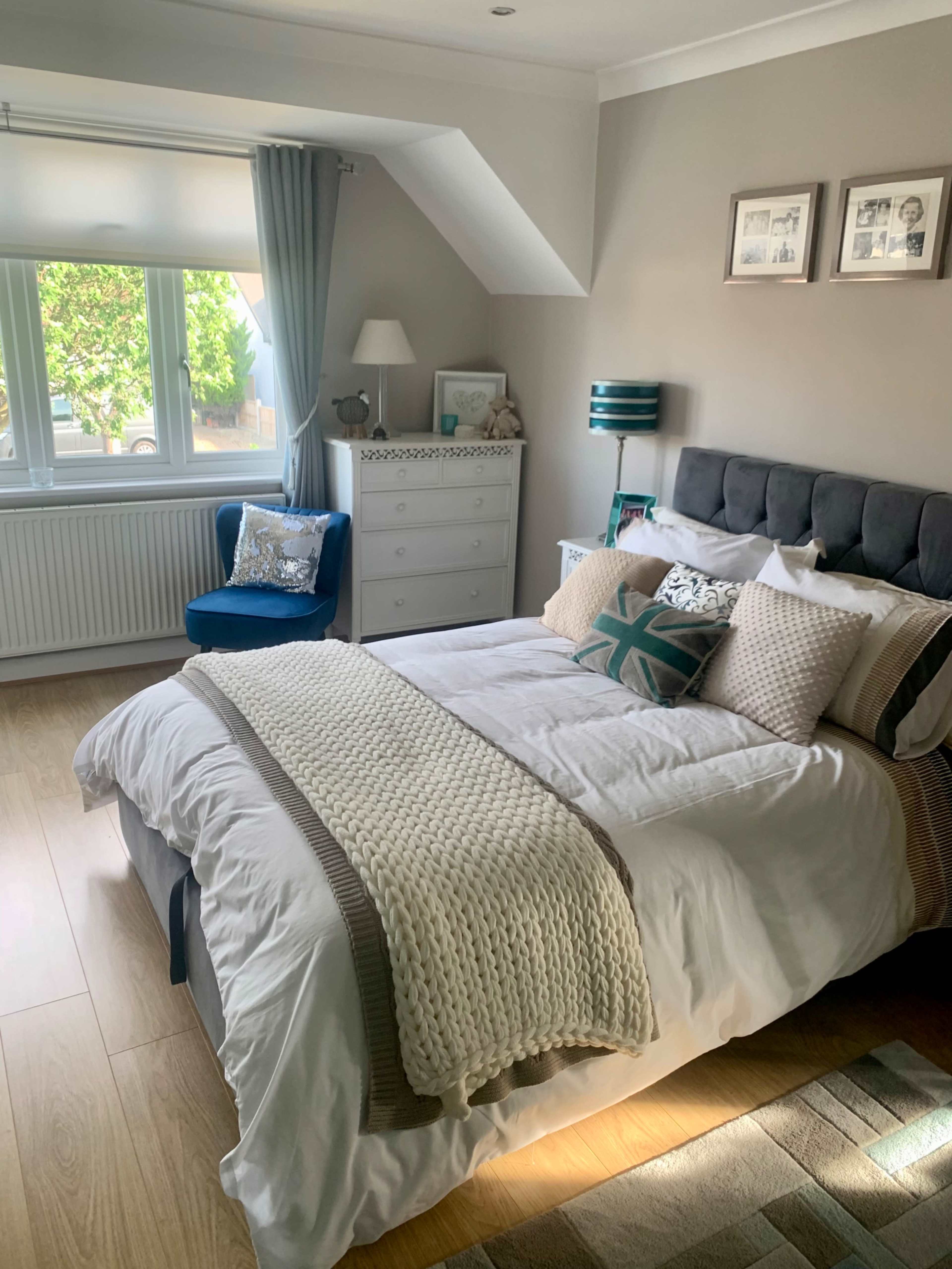 A neatly arranged bedroom with a double bed, decorative pillows, a dresser, and a cozy armchair beside a large window.