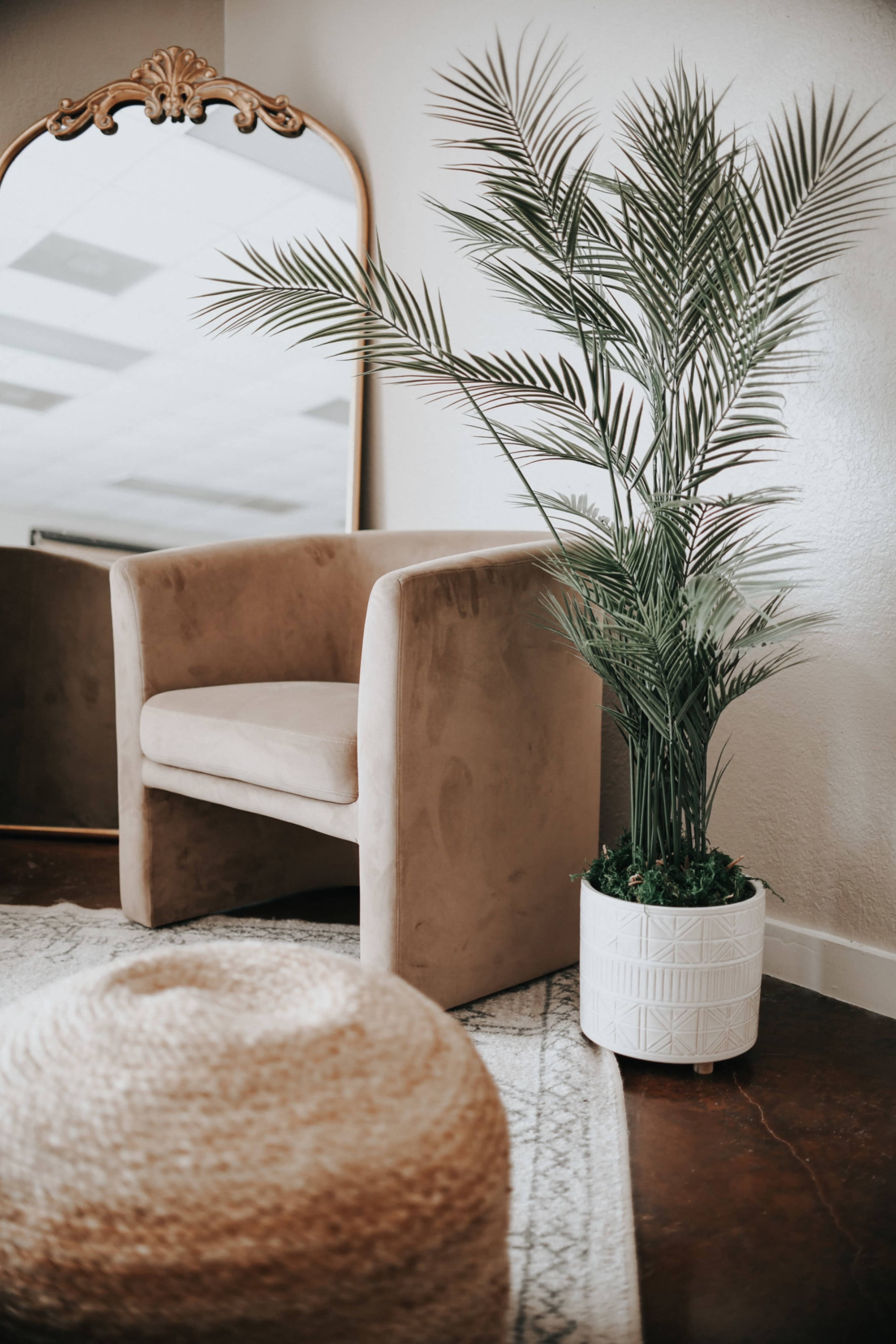 A beige upholstered chair sits next to a large potted palm plant and a round woven pouf, with a mirror reflecting the scene in the background.