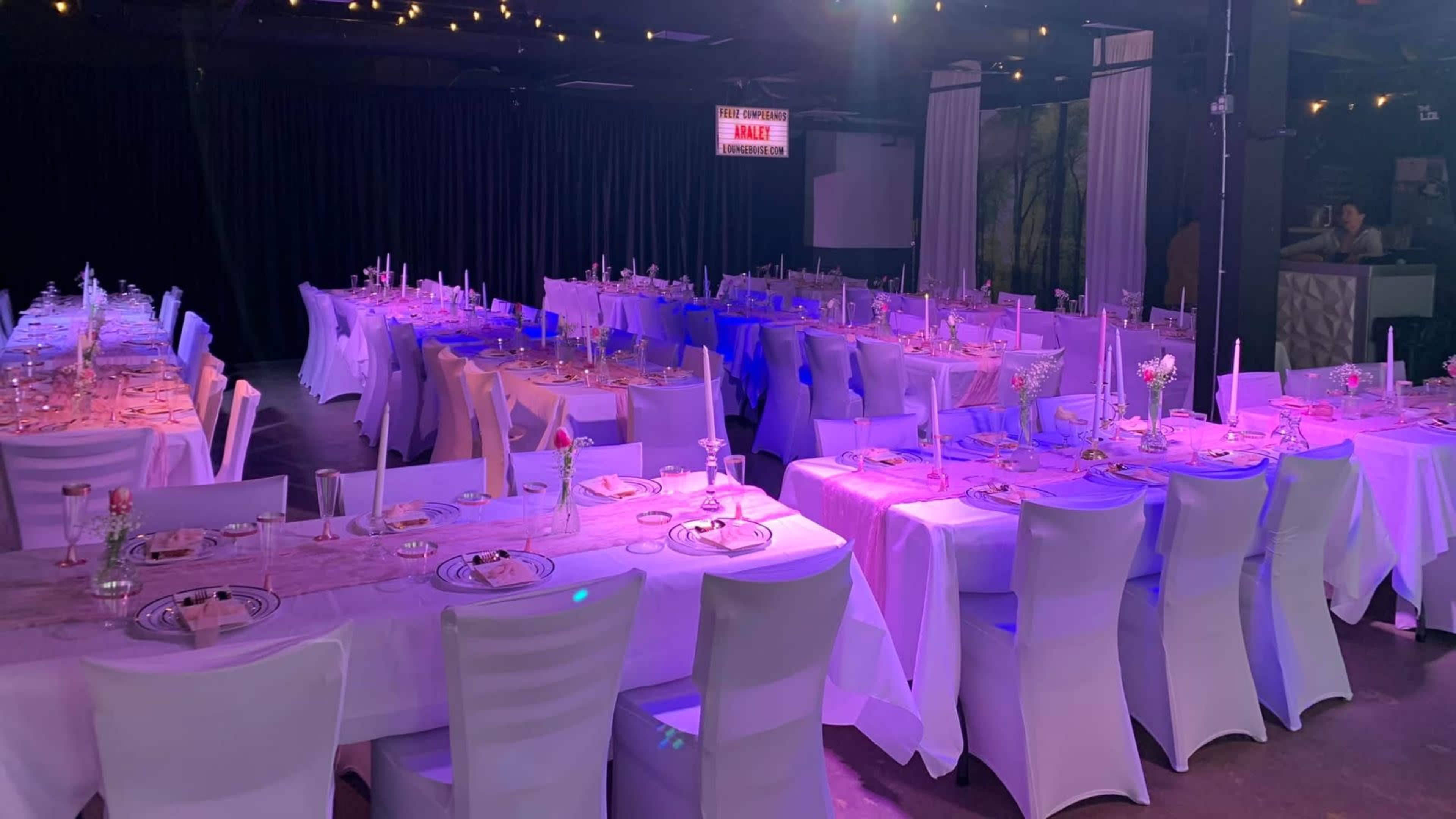 The image shows a spacious banquet hall set with rows of elegantly arranged tables covered in white tablecloths and decorated with centerpieces, ready for an event.