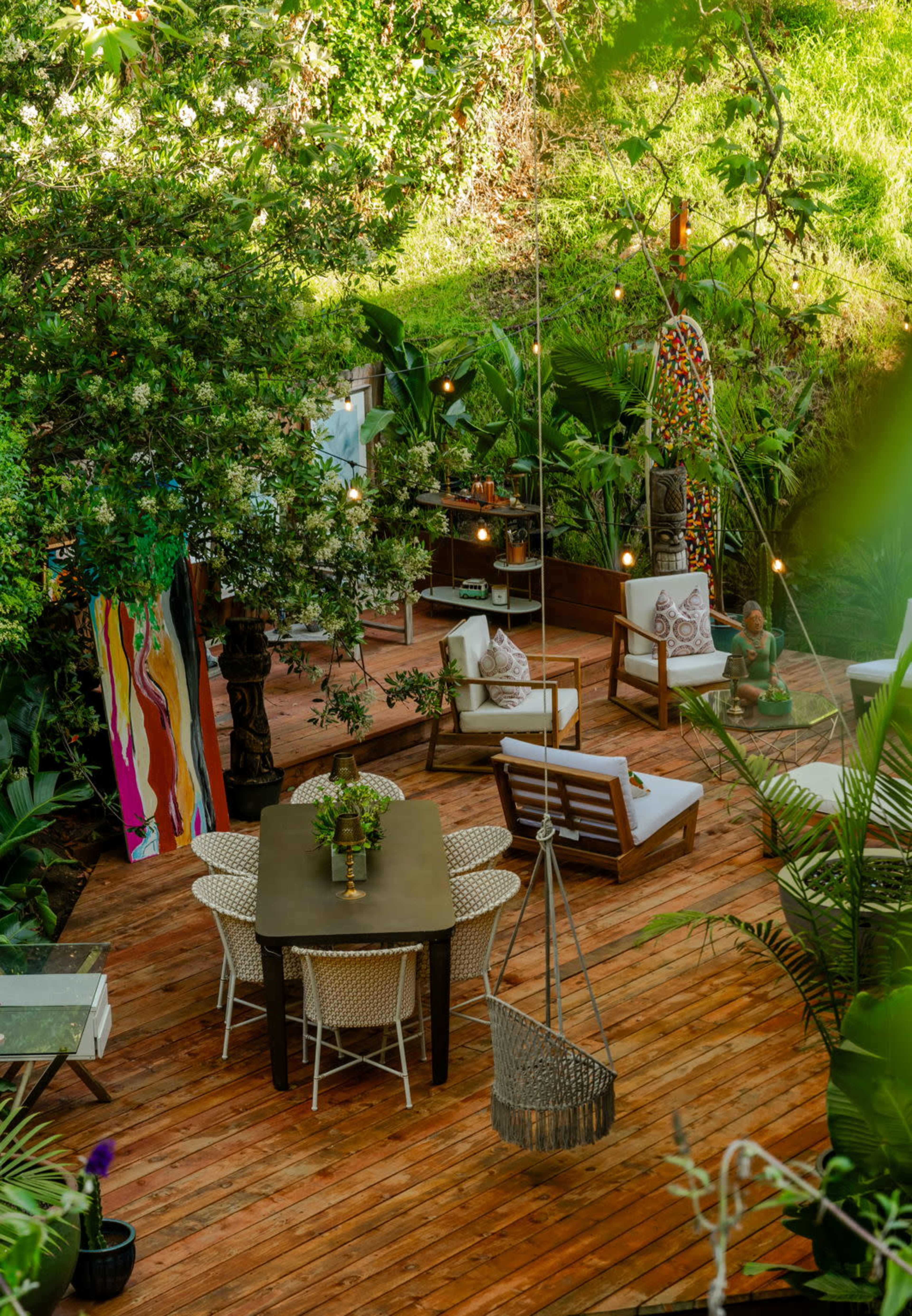 A wooden deck surrounded by lush greenery, featuring a dining table, chairs, and decorative elements like sculptures and colorful artwork.