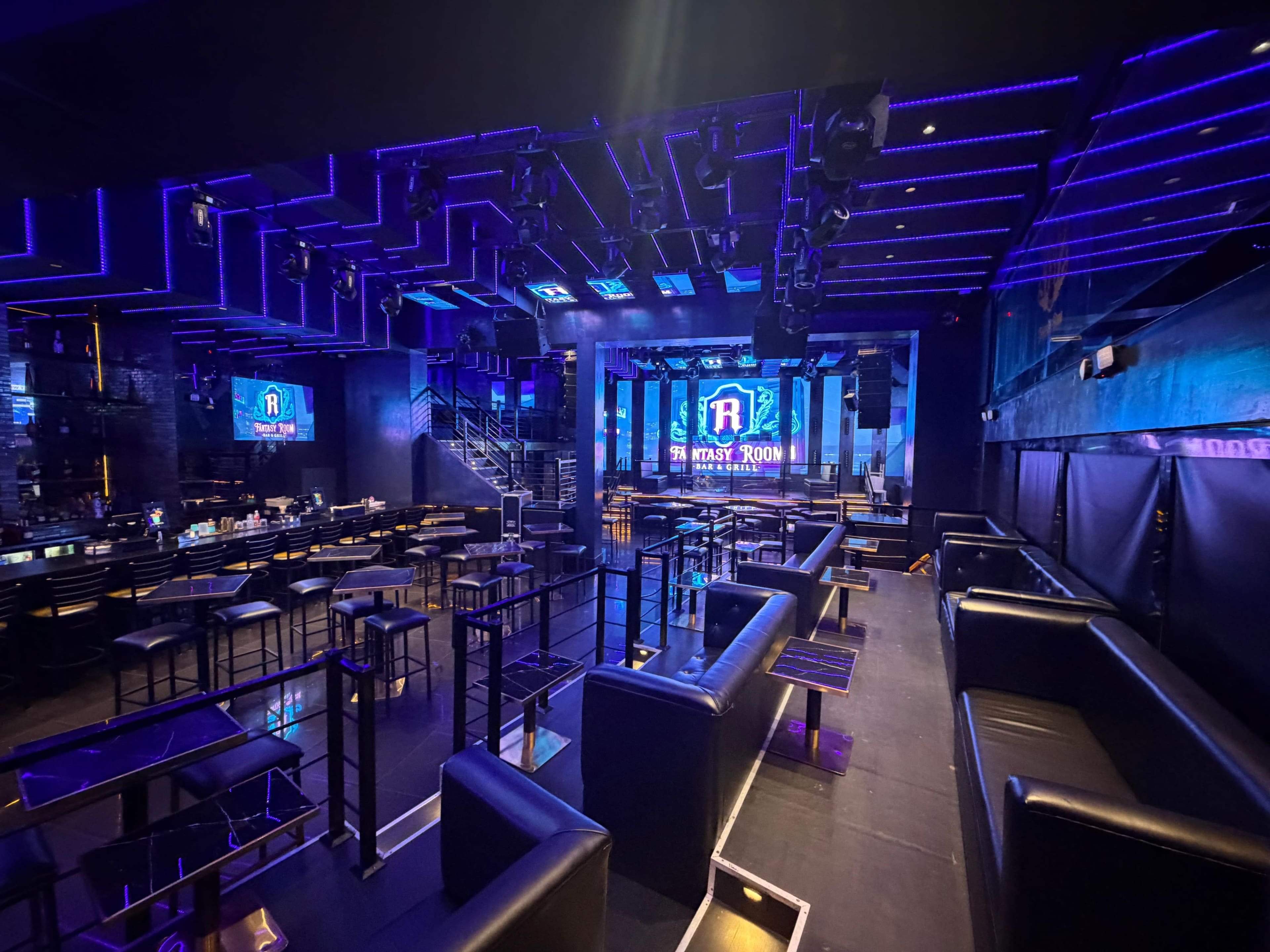 The image shows a dimly lit nightclub interior featuring black seating, a stage with lighting fixtures, and a large screen displaying the club's name.