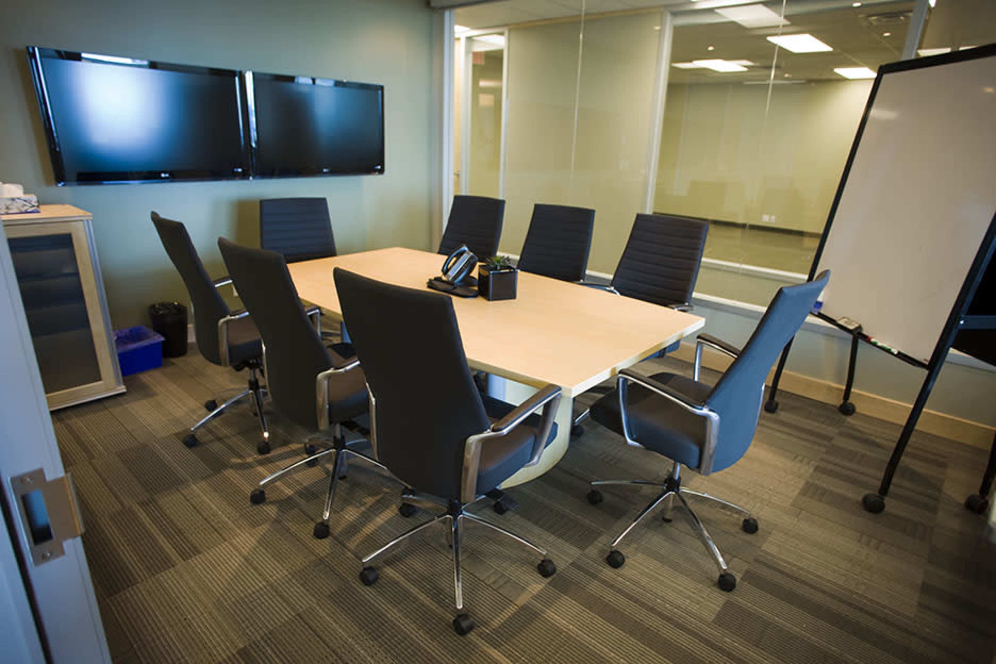 A conference room features a large table surrounded by eight ergonomic chairs, with two wall-mounted screens, a whiteboard, and a phone on the table.