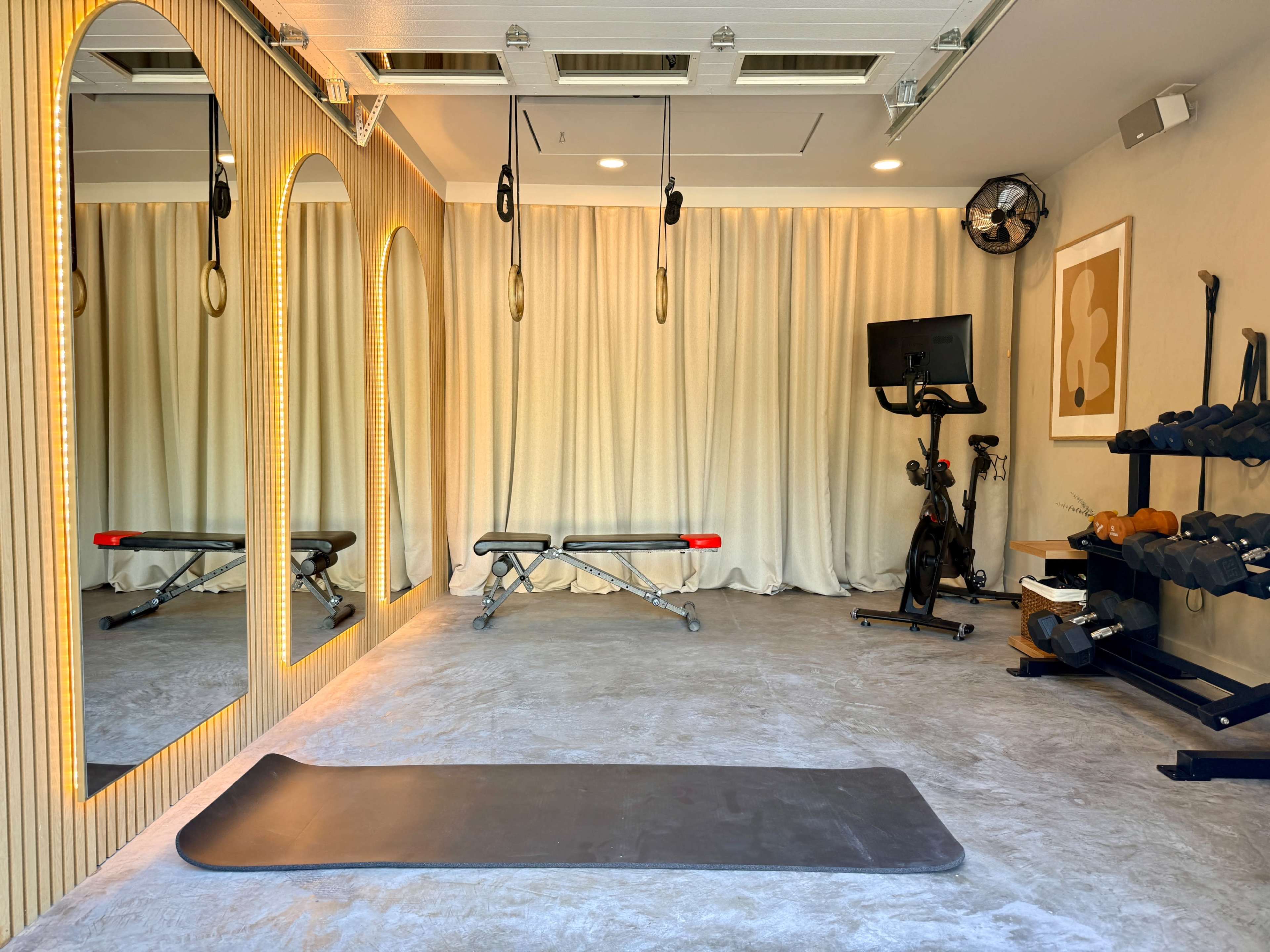 The image shows a home gym featuring mirrors, exercise equipment, and a mat on a concrete floor.