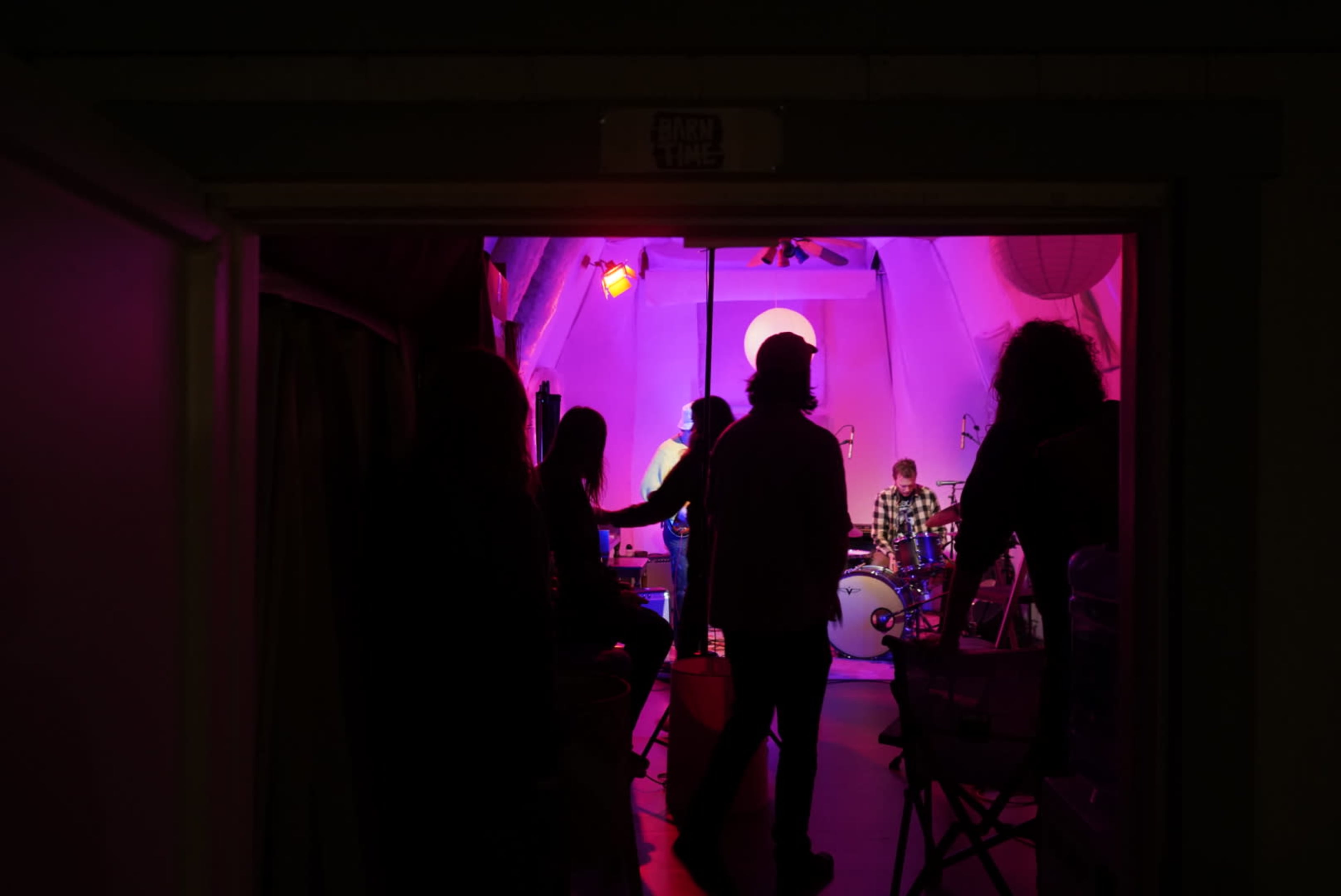 A group of silhouetted people watches a band perform under colored stage lighting in a small venue.