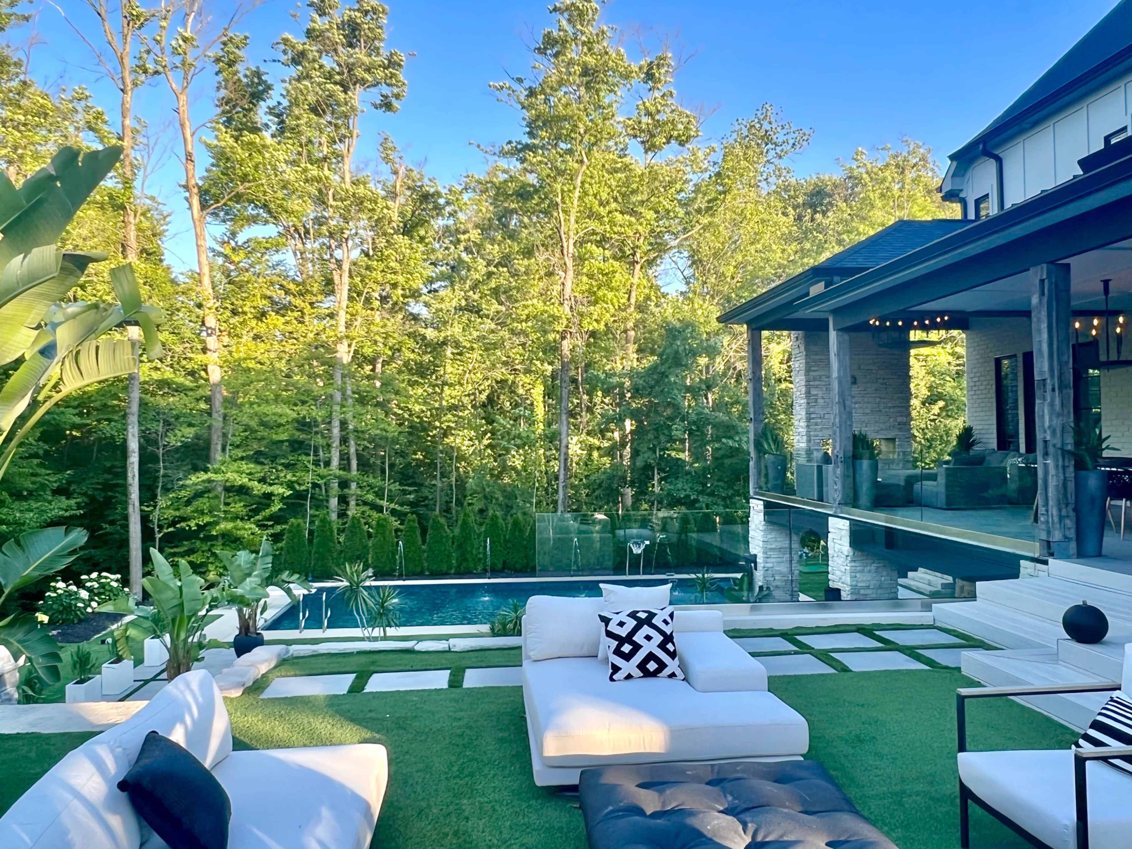 A spacious outdoor area features modern furniture set on green grass near a pool surrounded by trees and a contemporary house.