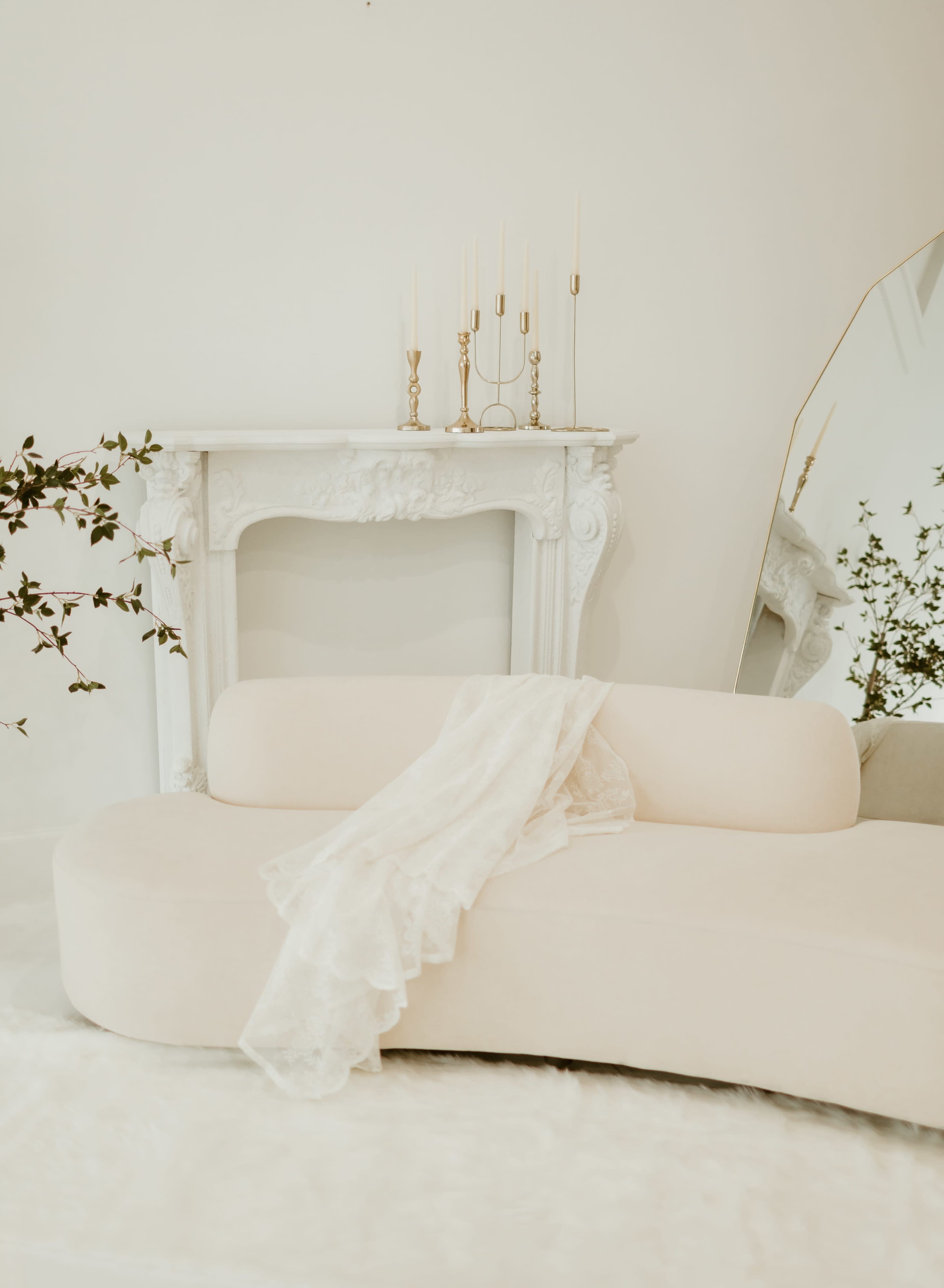A curved, light-colored sofa is positioned in front of a white fireplace adorned with candles, while a mirror reflects the scene.
