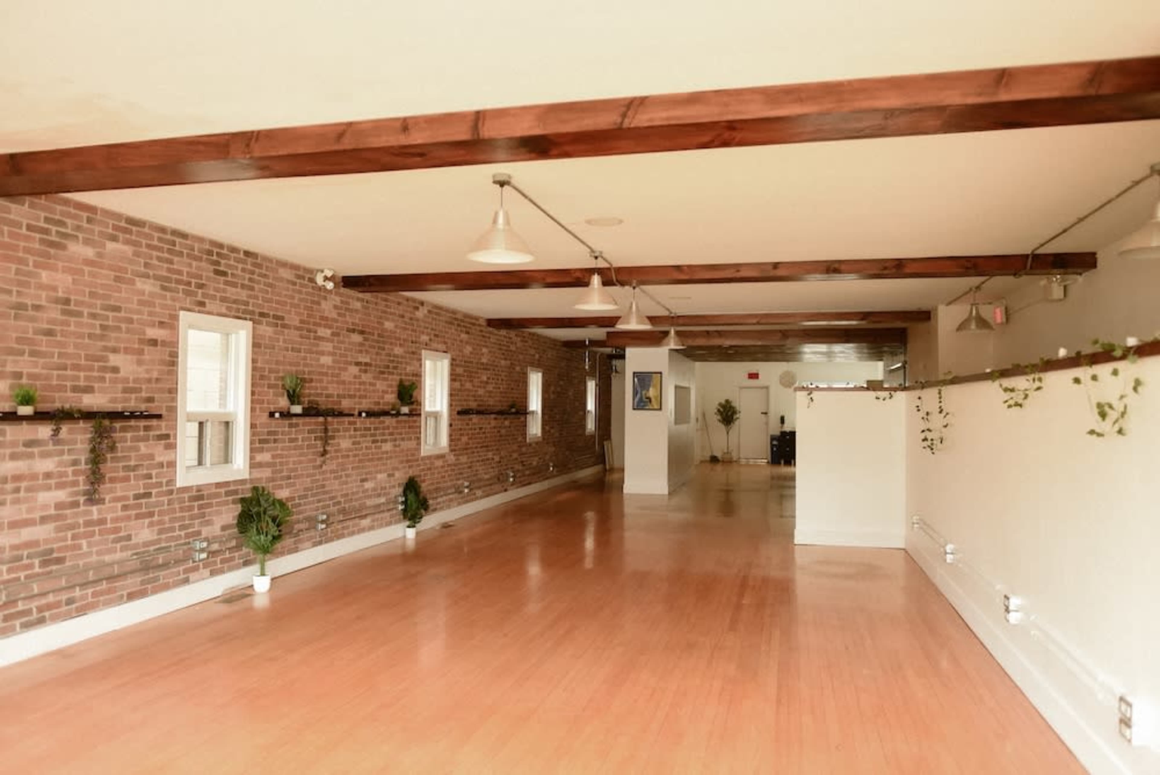 The image shows an empty room with wooden beams, a brick wall, potted plants on shelves, and smooth wooden flooring.