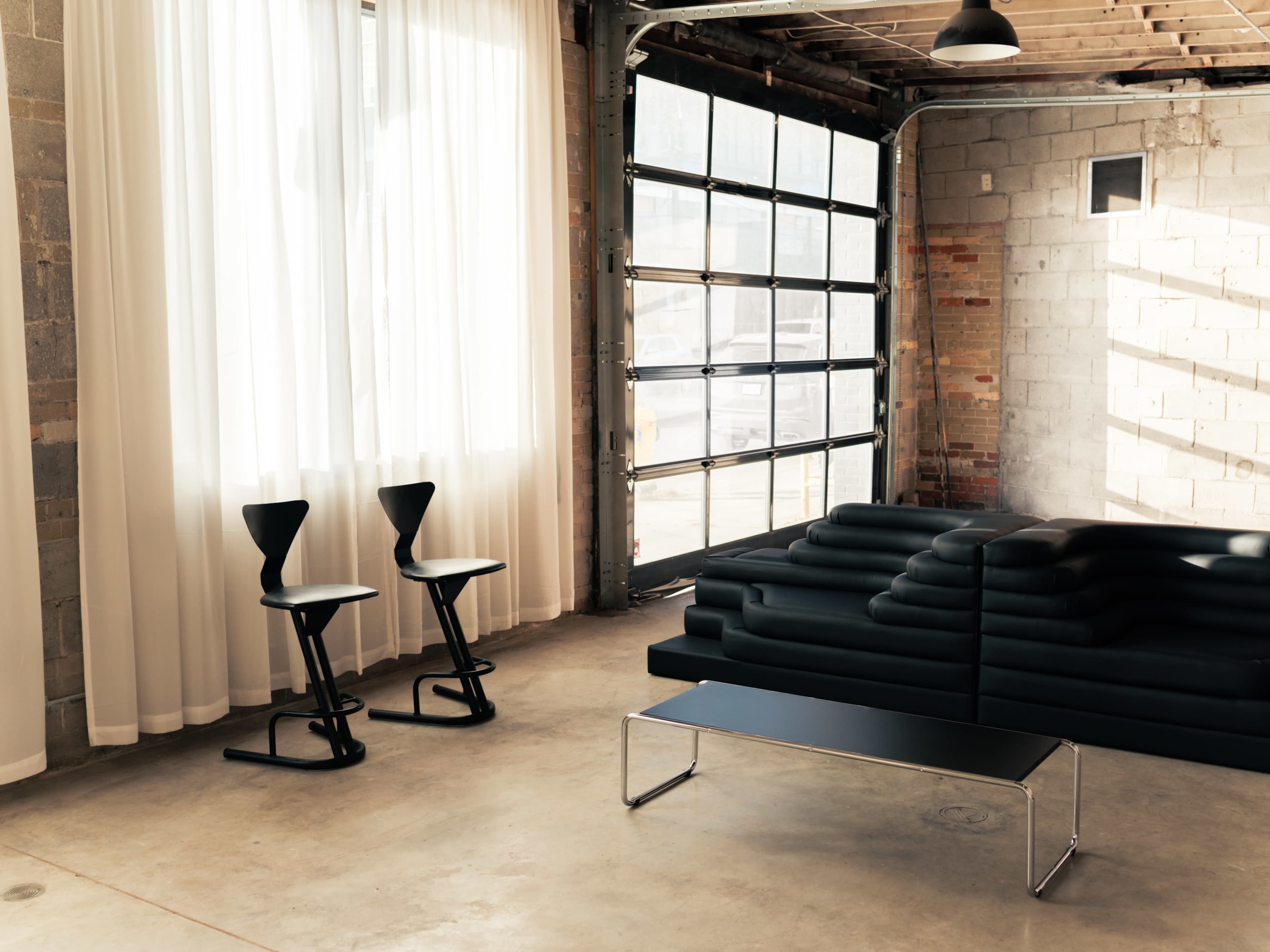The scene features a minimalist interior with a black sofa, two black chairs, a metal coffee table, large windows with white curtains, and exposed brick walls.