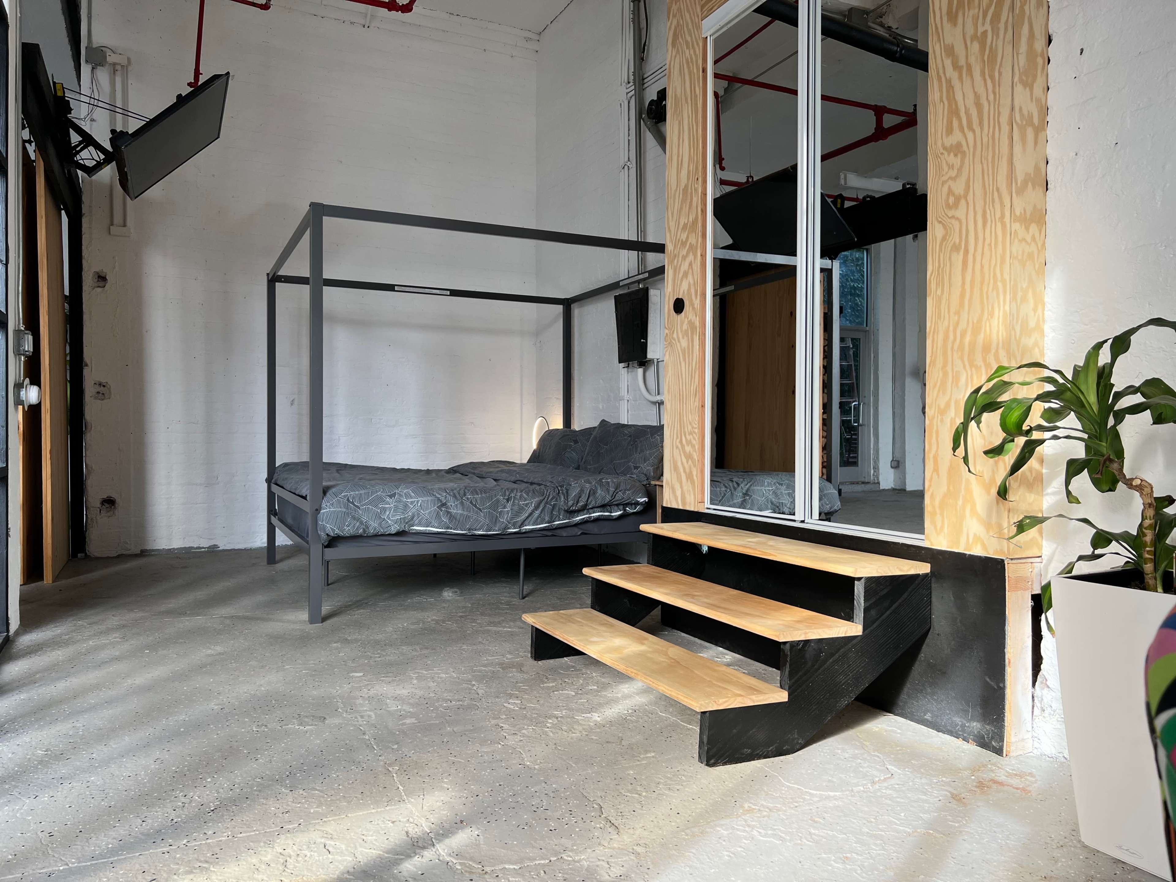 A minimalist room featuring a four-poster bed, a set of wooden stairs, and a large mirror against a wall.
