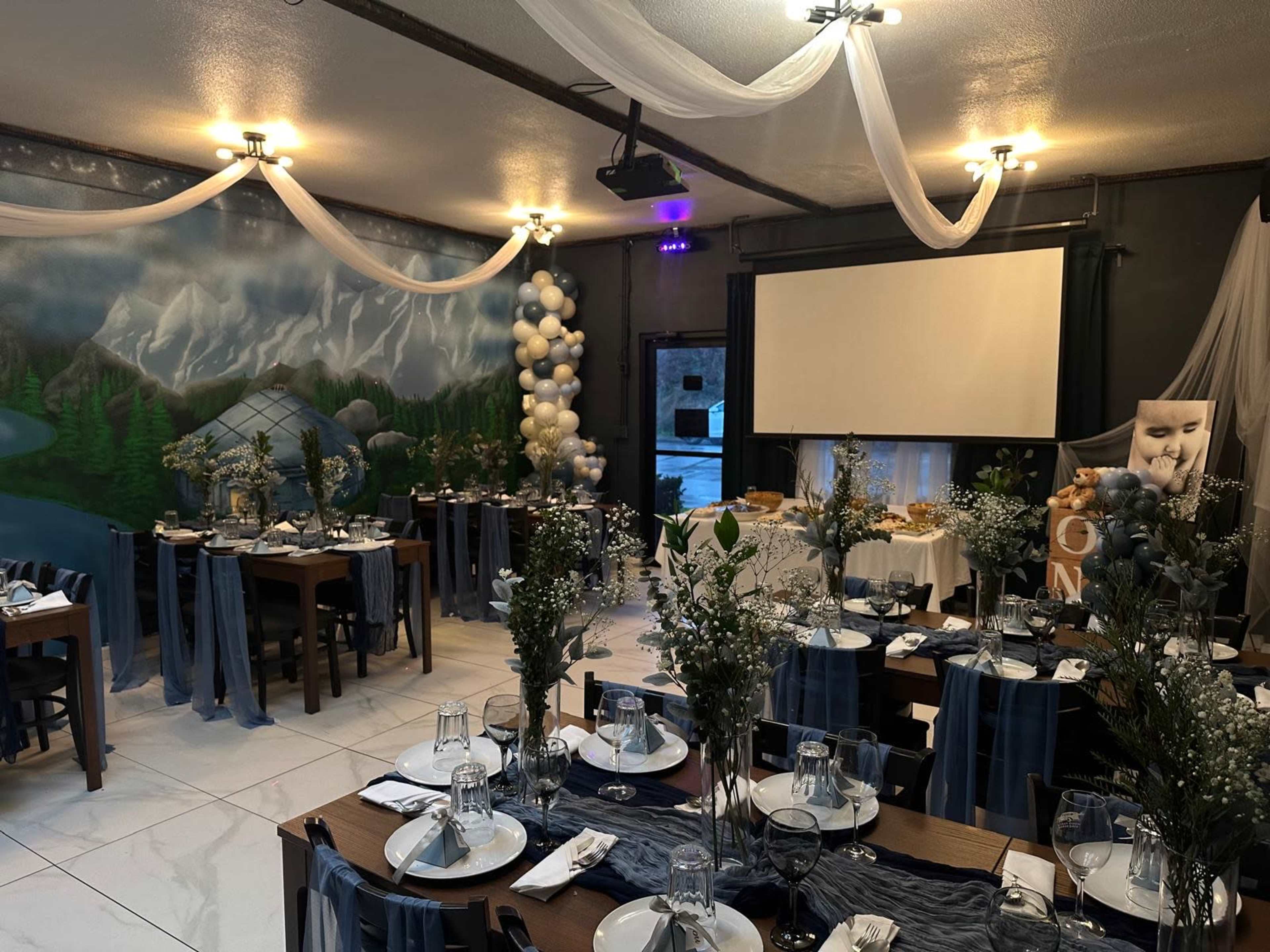 The image depicts a decorated event space featuring long tables set with dishware, greenery, and white floral arrangements, surrounded by a mural of mountains and a large projection screen.