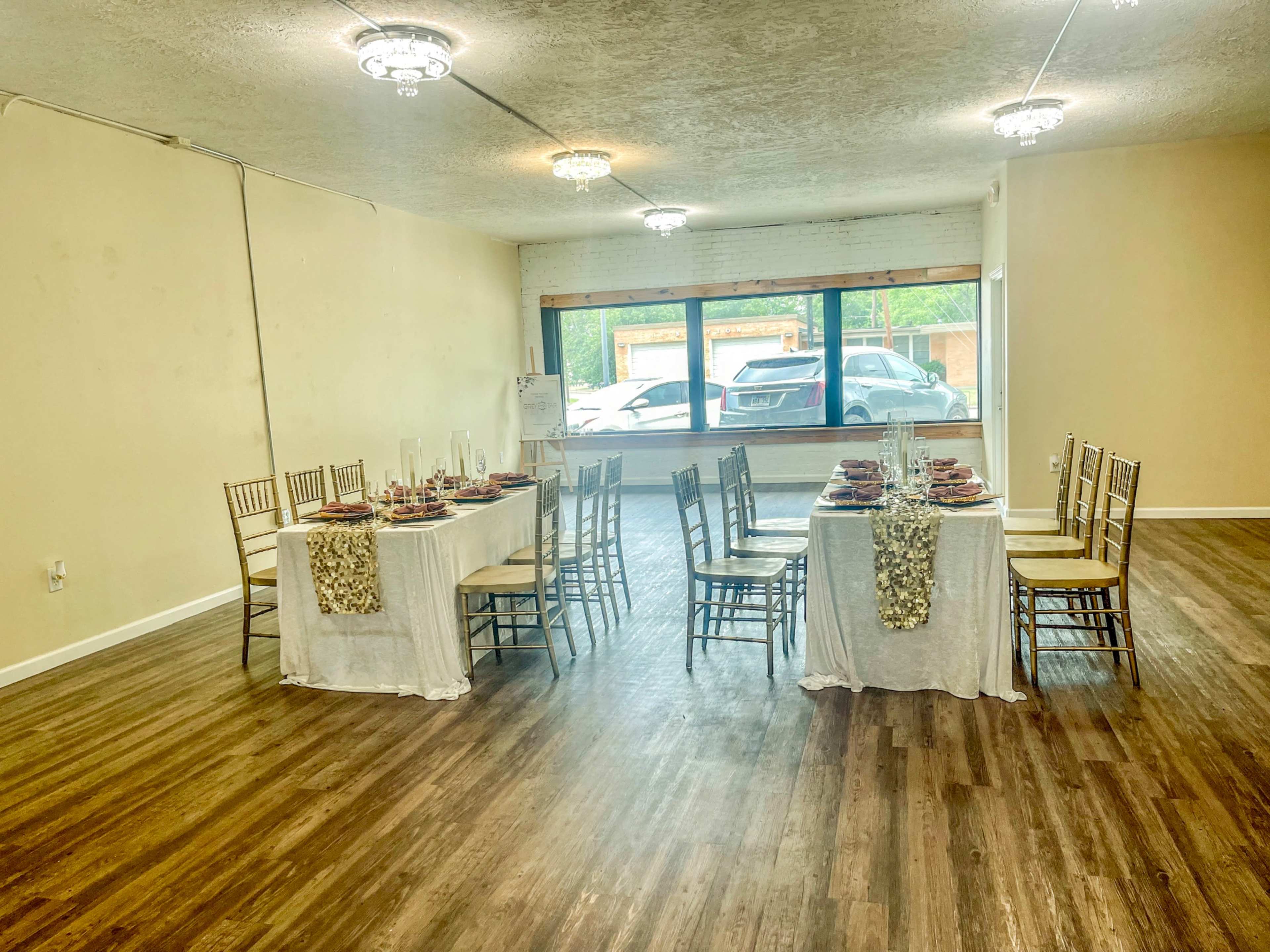 An elegantly set dining area featuring two tables with white tablecloths, arranged in a well-lit room with wooden floors and large windows.