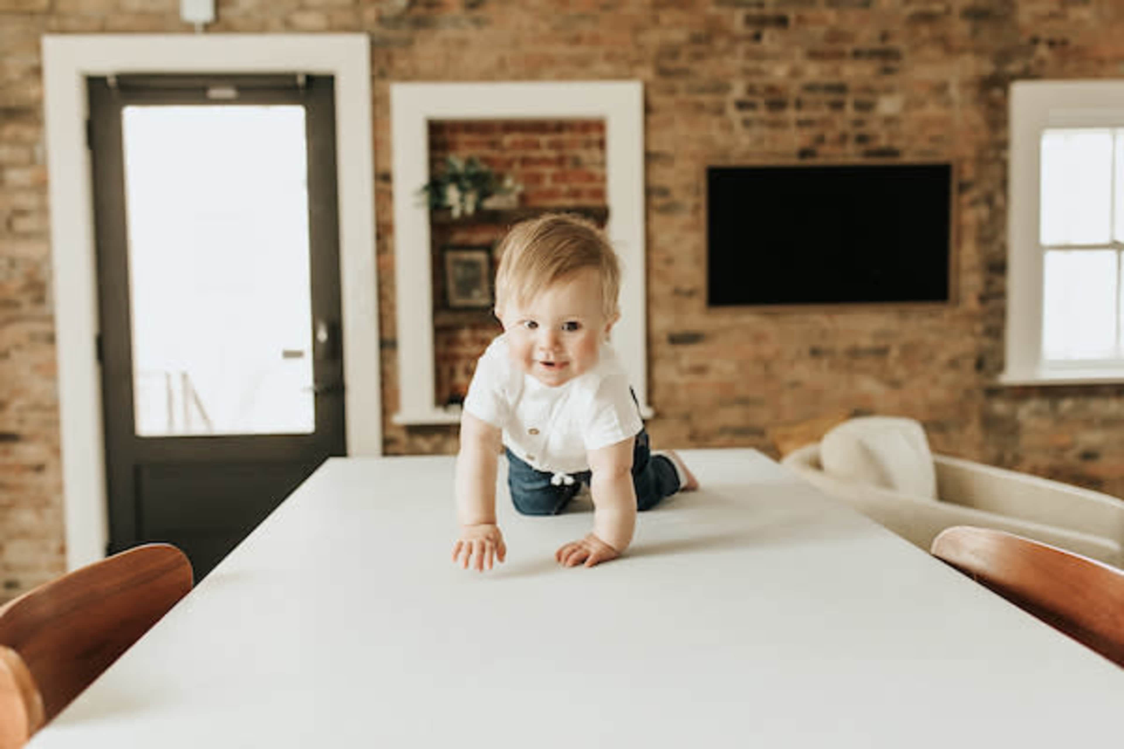 A baby is crawling on a white table in a room with exposed brick walls and a large window.
