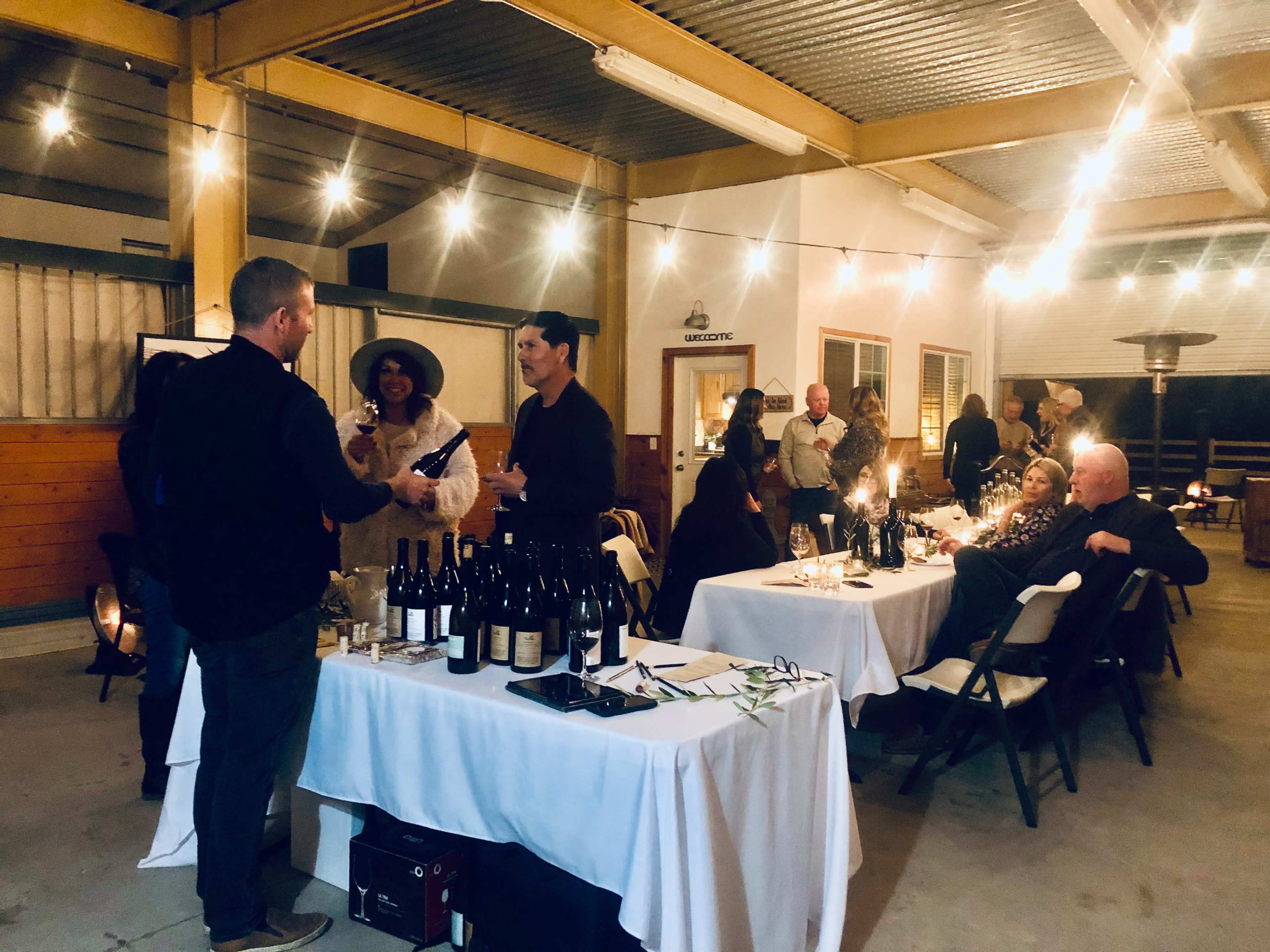 A gathering is taking place indoors with people socializing, tables set with wine bottles, and string lights overhead.