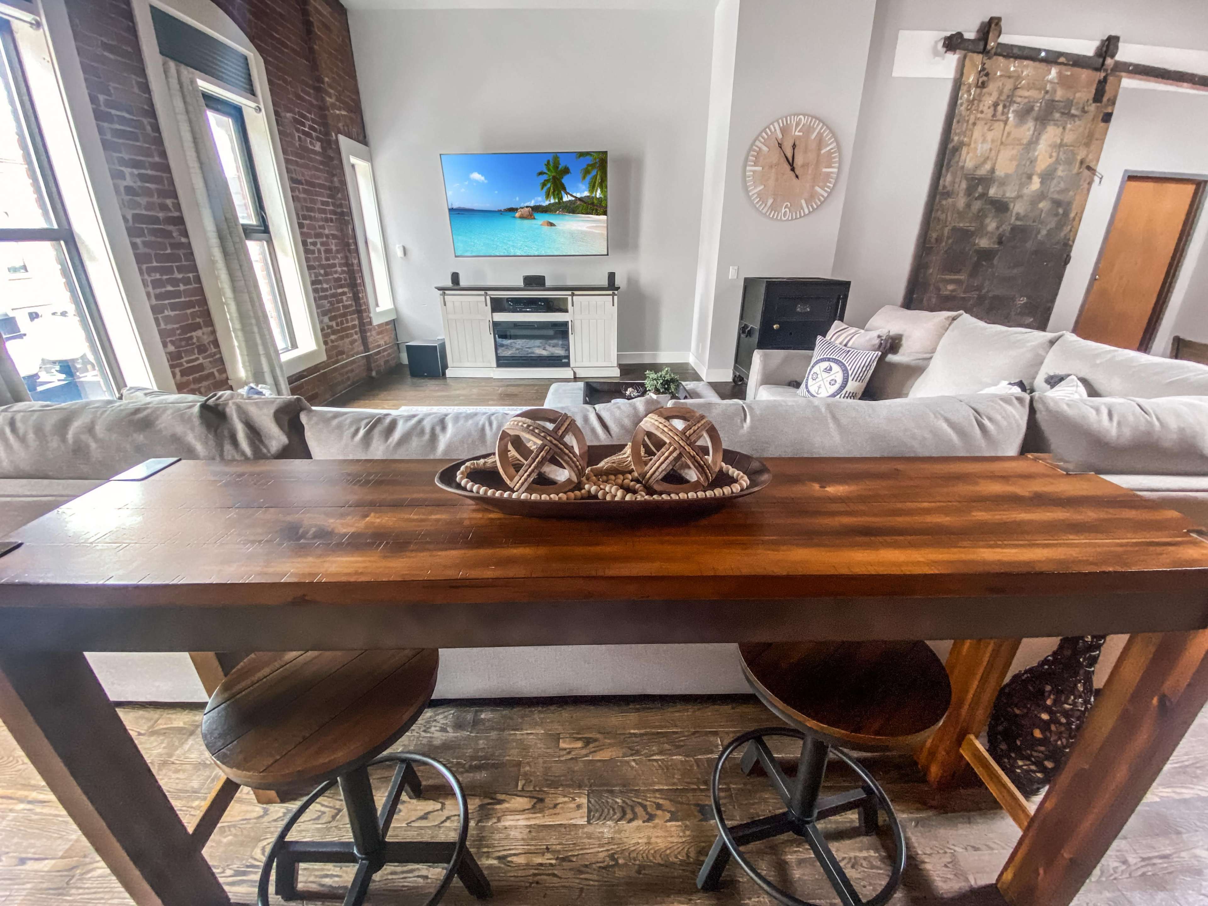 A rustic wooden table with two stools is placed in front of a living room featuring a television, a beach scene on the screen, and an exposed brick wall.