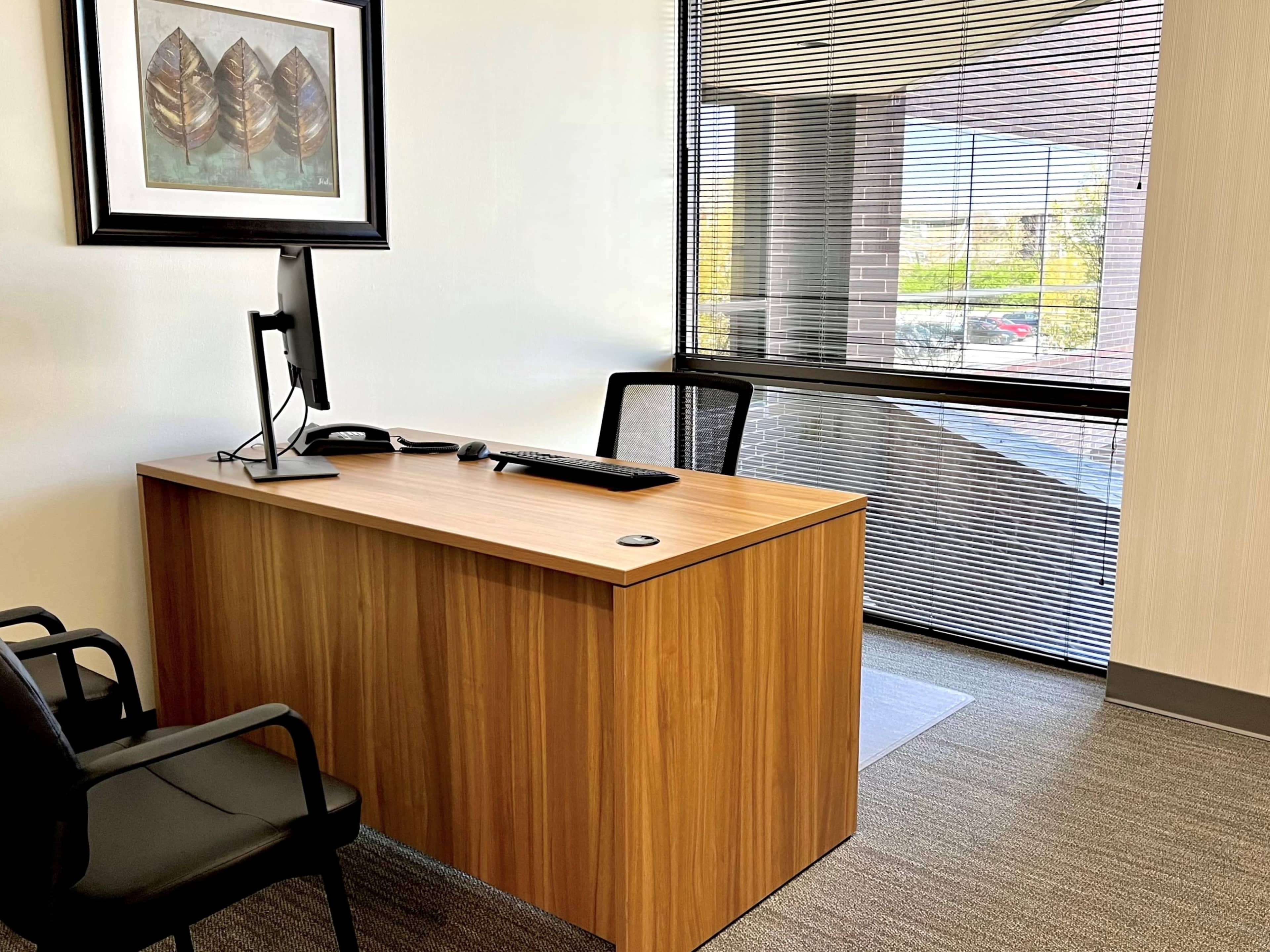 The image shows a minimalist office interior featuring a wooden desk, a computer monitor, and a chair next to a large window with blinds.