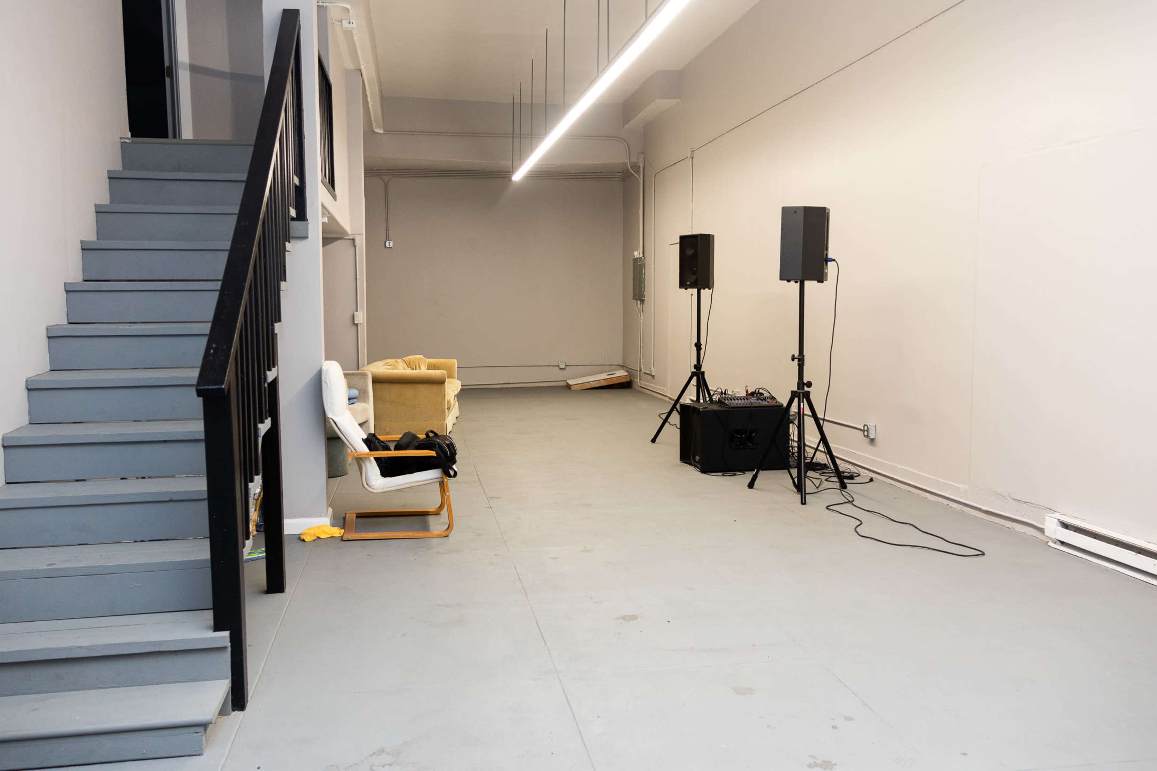 The image shows a spacious, empty room with a staircase on the left, a yellow armchair and speakers positioned along the right wall.