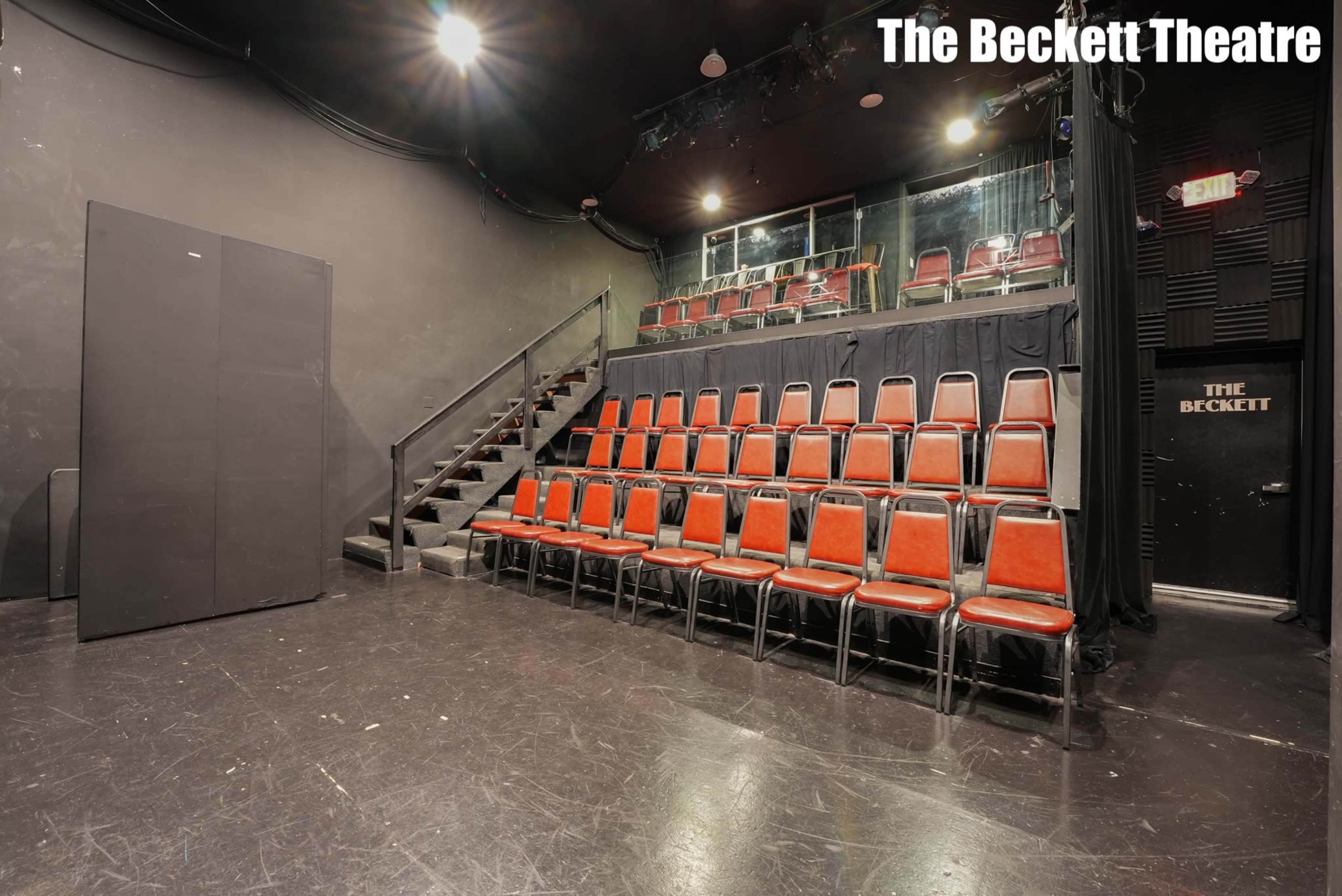 The image shows a small theater space with black walls, a staircase leading to a raised seating area, and rows of red chairs arranged in front of it.