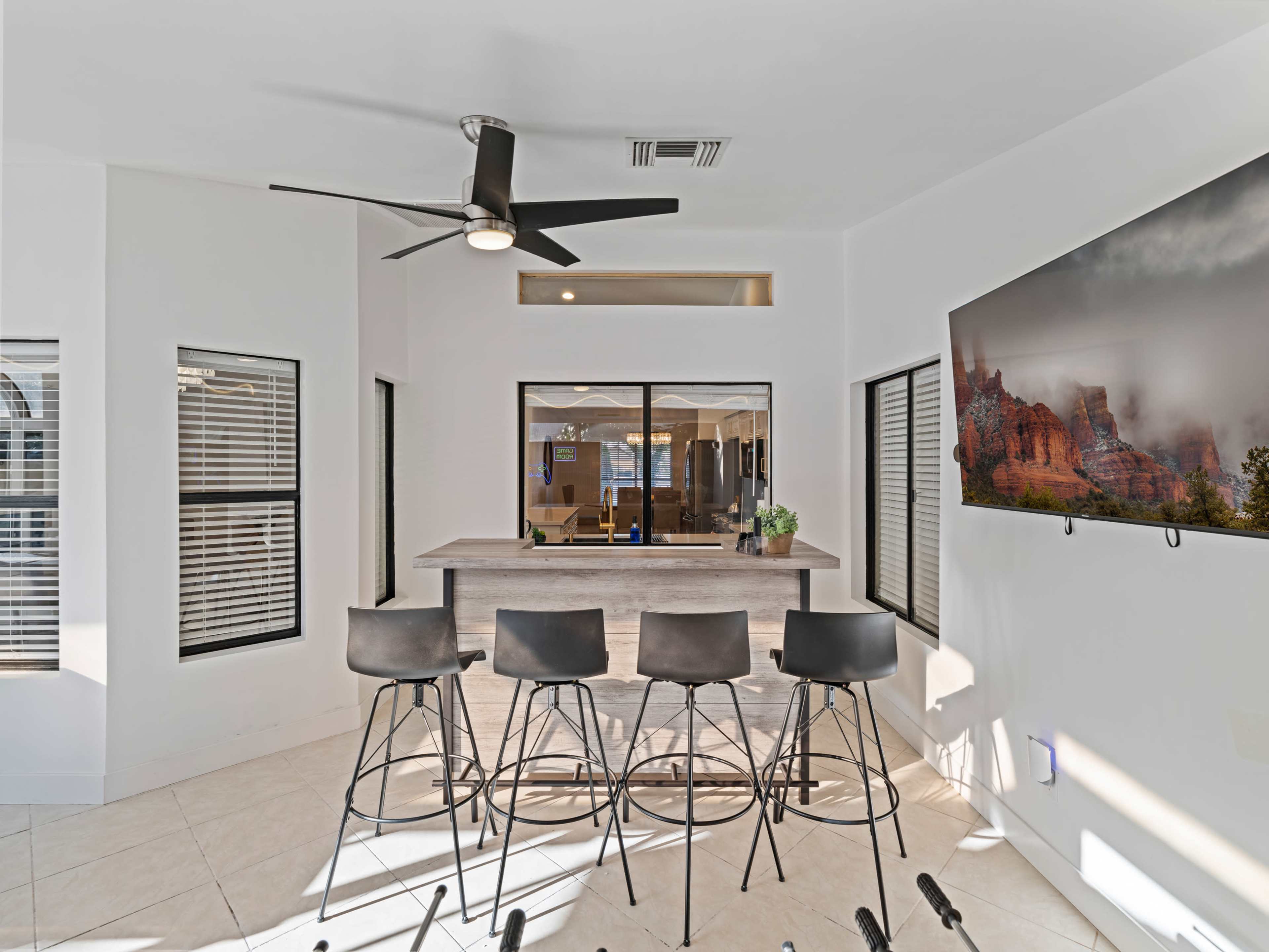 A modern indoor bar area with high stools, a ceiling fan, large windows, and a mounted TV on the wall.