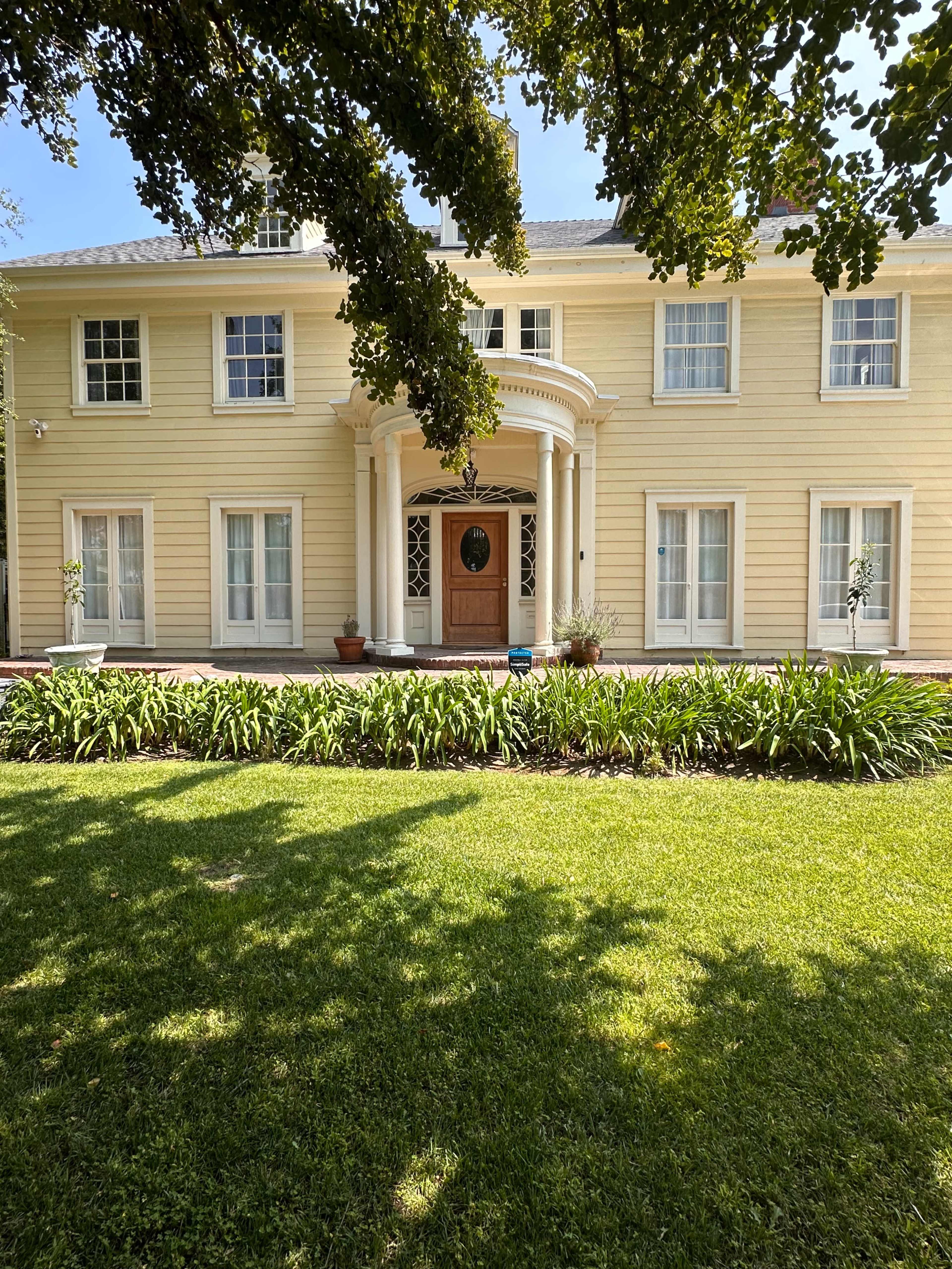 A two-story yellow house with white trim, featuring a central entrance with a decorative columned porch and a well-maintained garden in front.