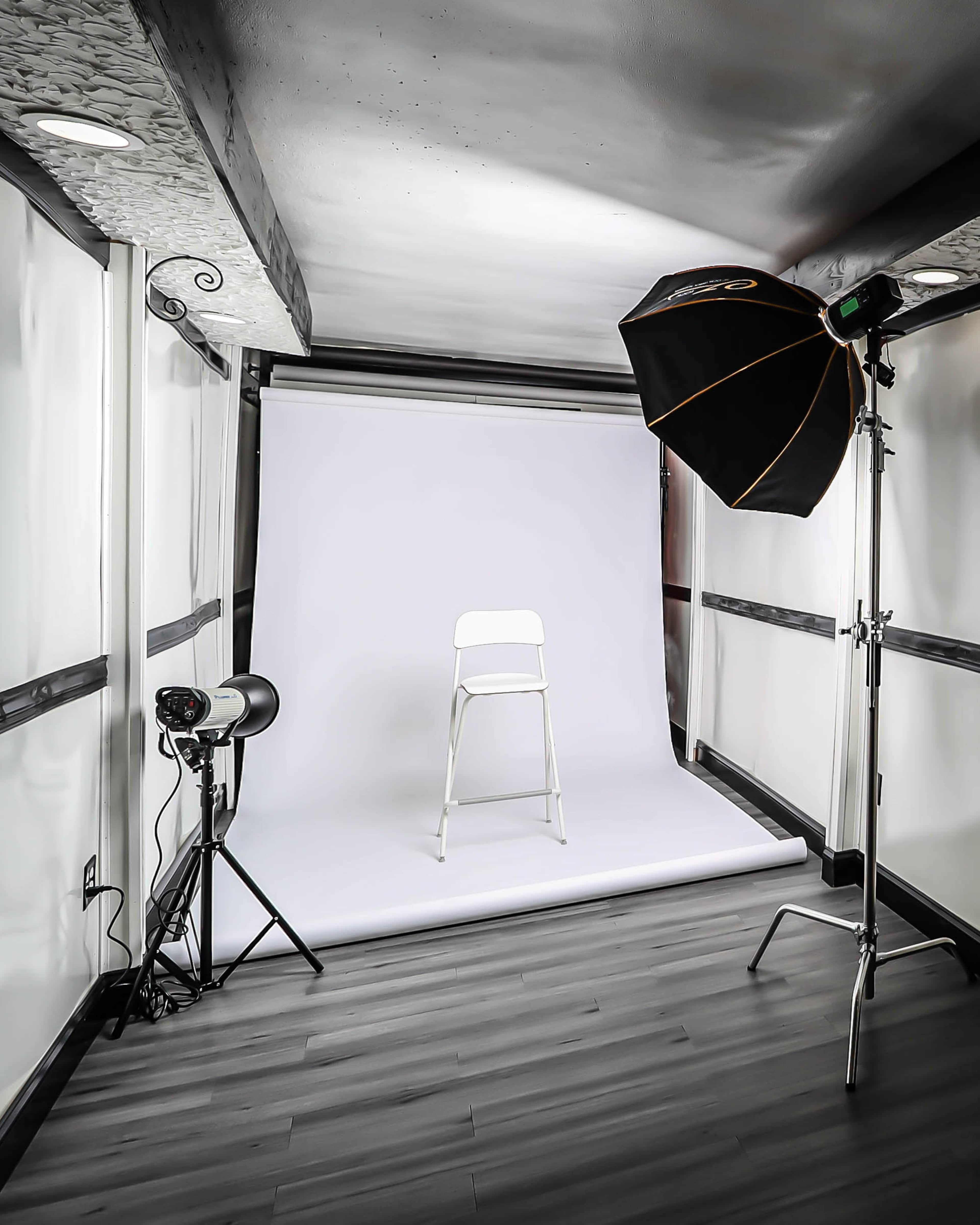 A photography studio features a white backdrop, a single white chair, and lighting equipment set up in a corner.