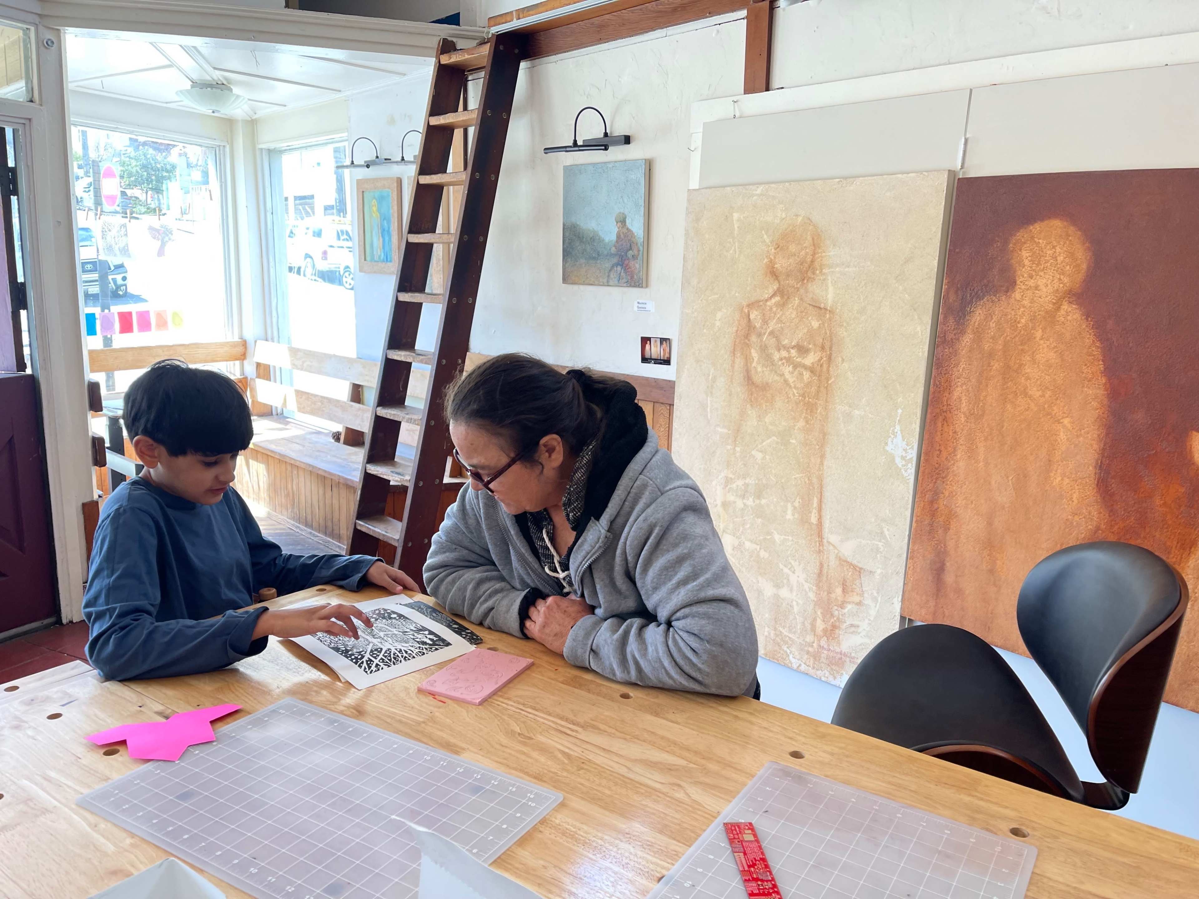 A child and an adult sit at a wooden table, examining a book, while artwork hangs on the walls of a well-lit space.