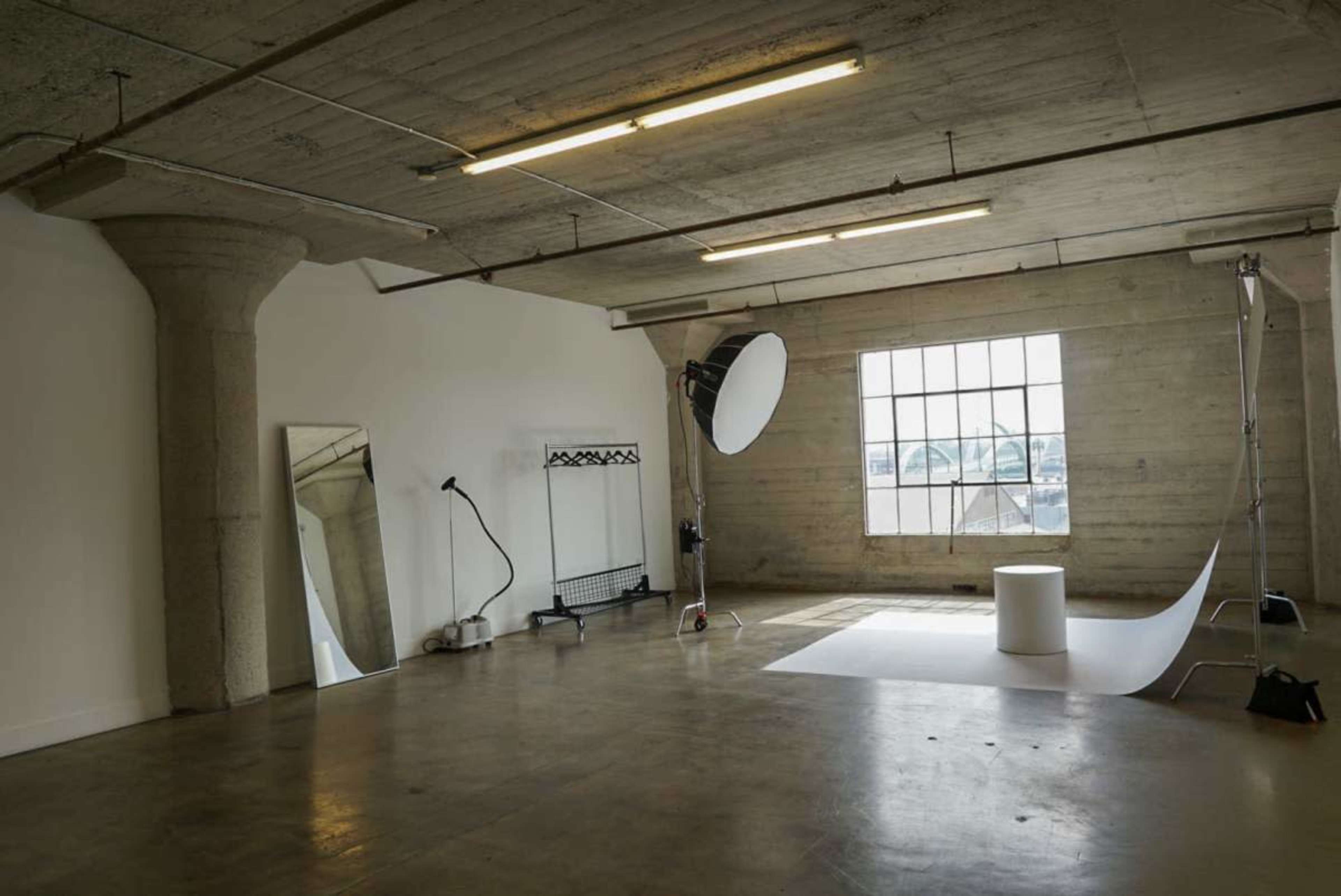 The image shows a spacious, empty studio with a large window, a backdrop setup, a mirror, and minimalist equipment.