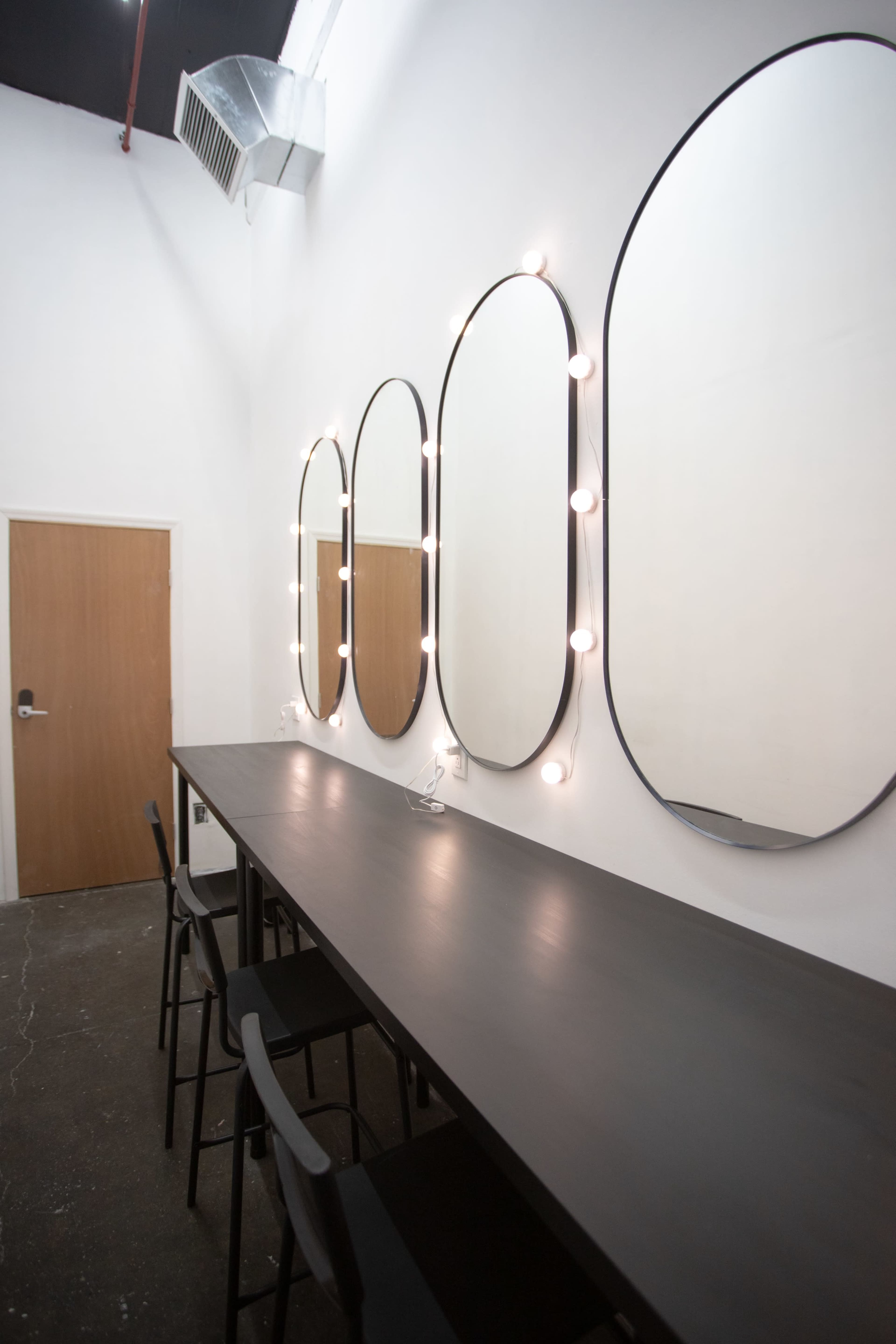 A simple room with four oval mirrors framed by light bulbs above a long, dark table and black chairs, with a door on the left.