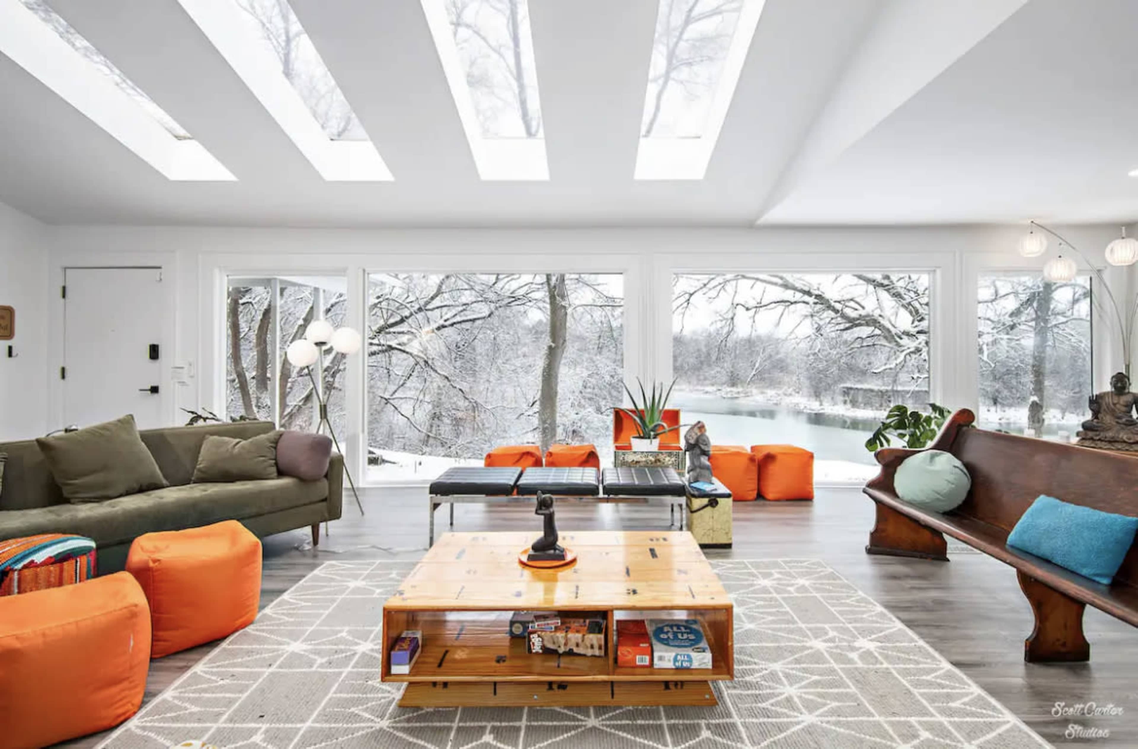 The living room features large windows overlooking a snowy landscape, with a mix of seating options including a green sofa, orange poufs, and a wooden bench, complemented by a wooden coffee table.