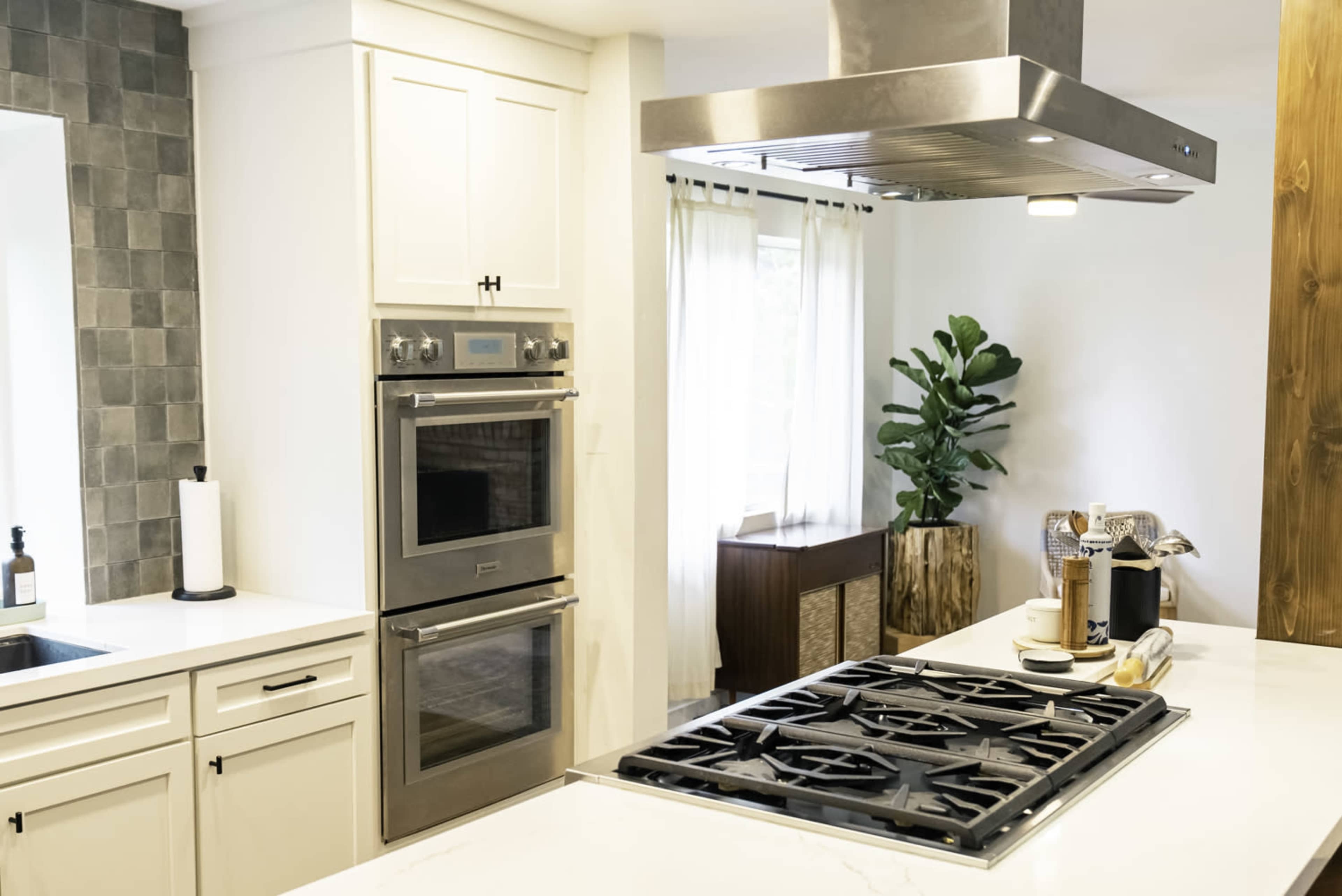 The image shows a modern kitchen featuring a gas cooktop, double wall ovens, a stainless steel range hood, and a window with sheer curtains.