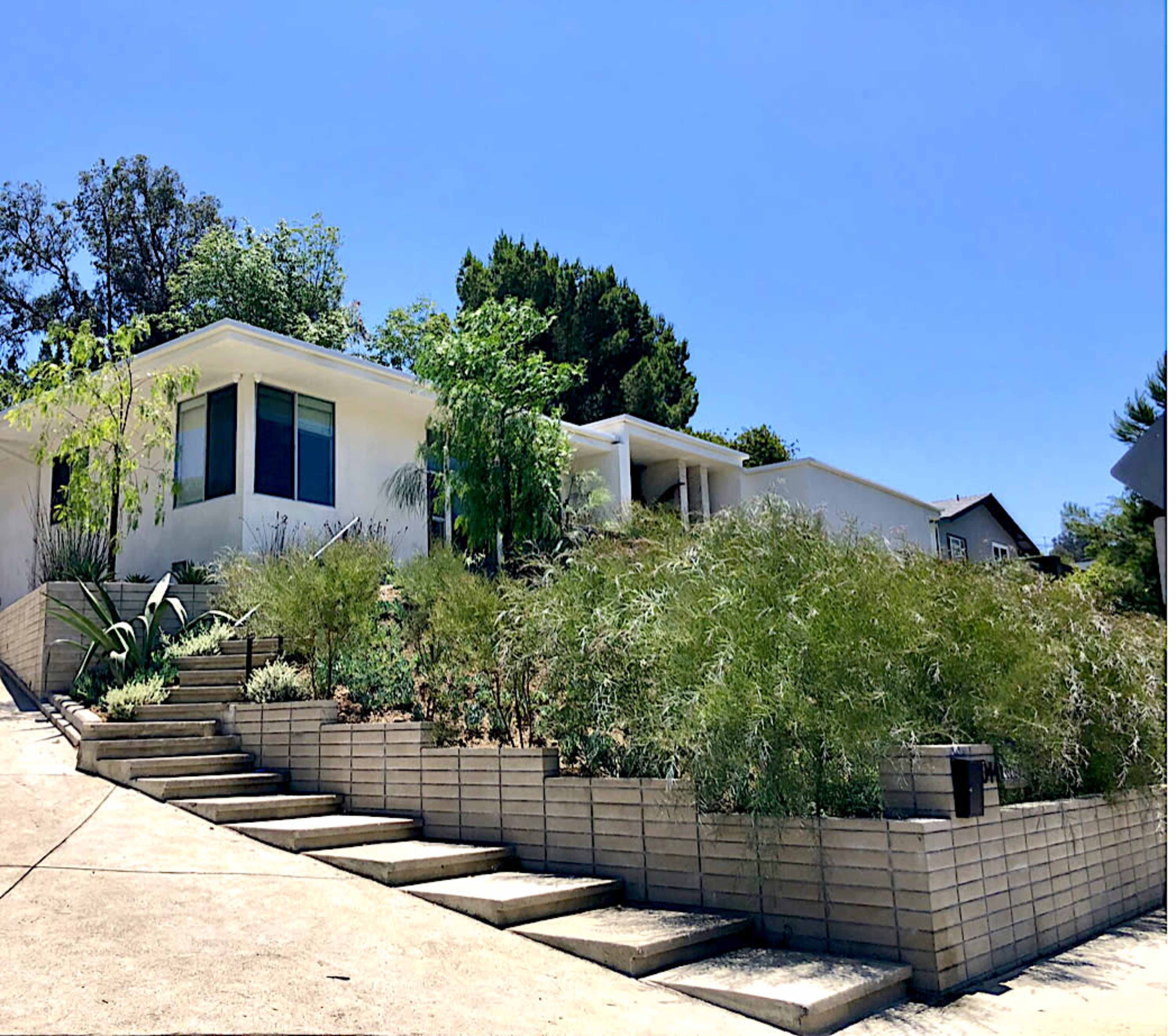 A modern house is situated on a hillside with a series of concrete steps leading up to it, surrounded by landscaped greenery.