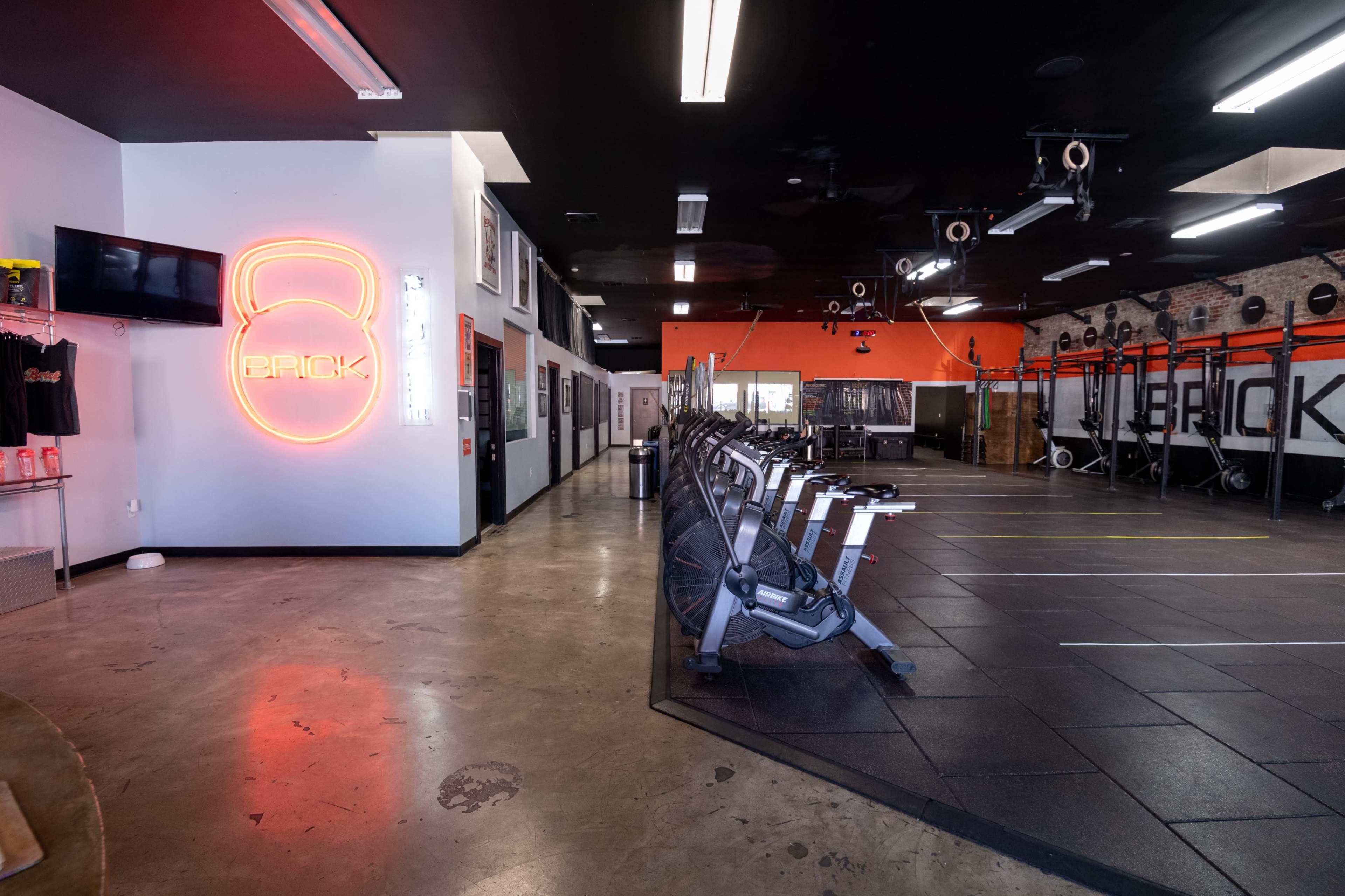 The image shows a fitness studio featuring rowers arranged in rows, with a neon sign displaying "PACK" on the wall and black rubber flooring.