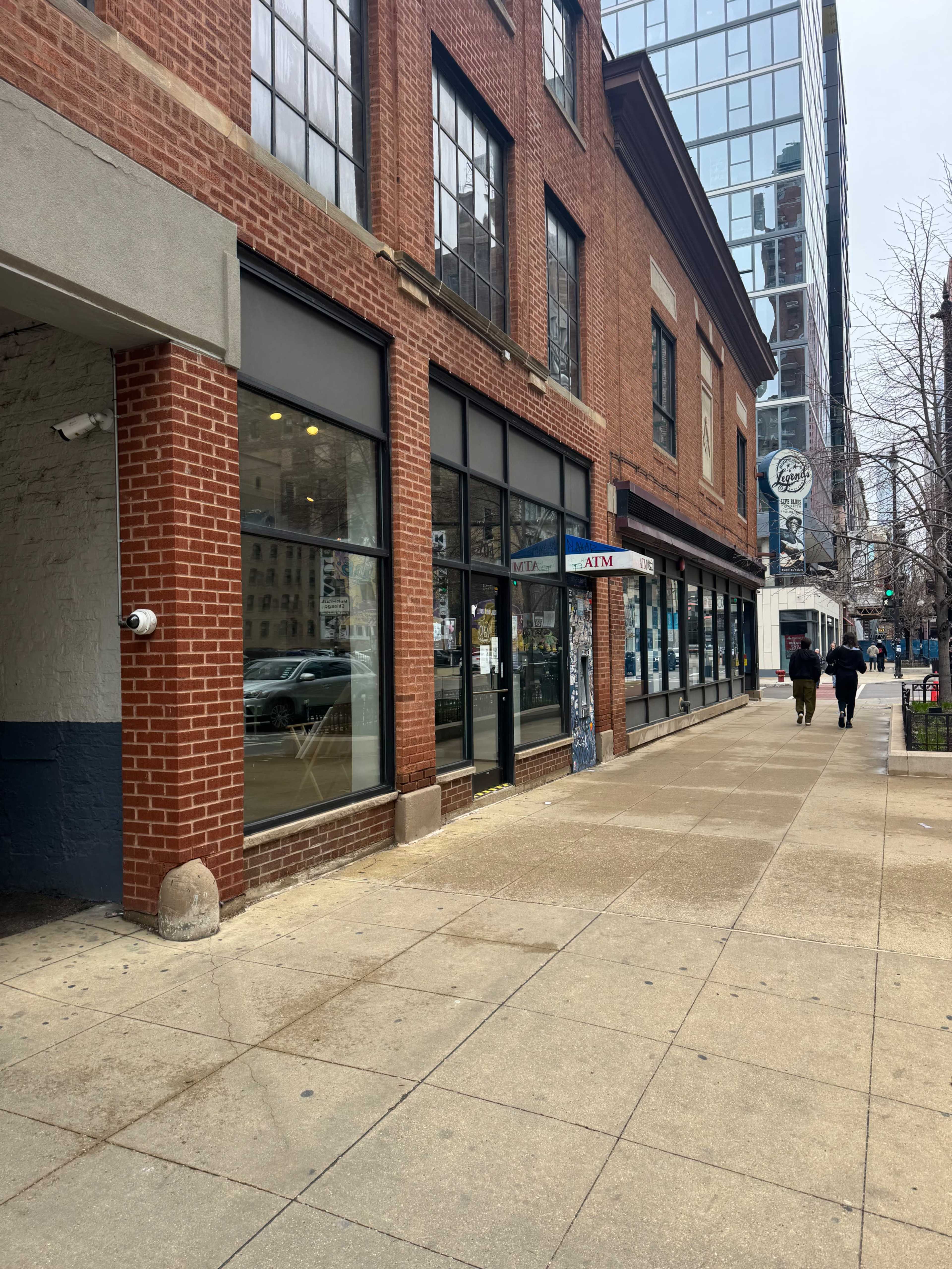 The image shows a brick building with large windows on a city sidewalk, alongside a street with pedestrians walking.