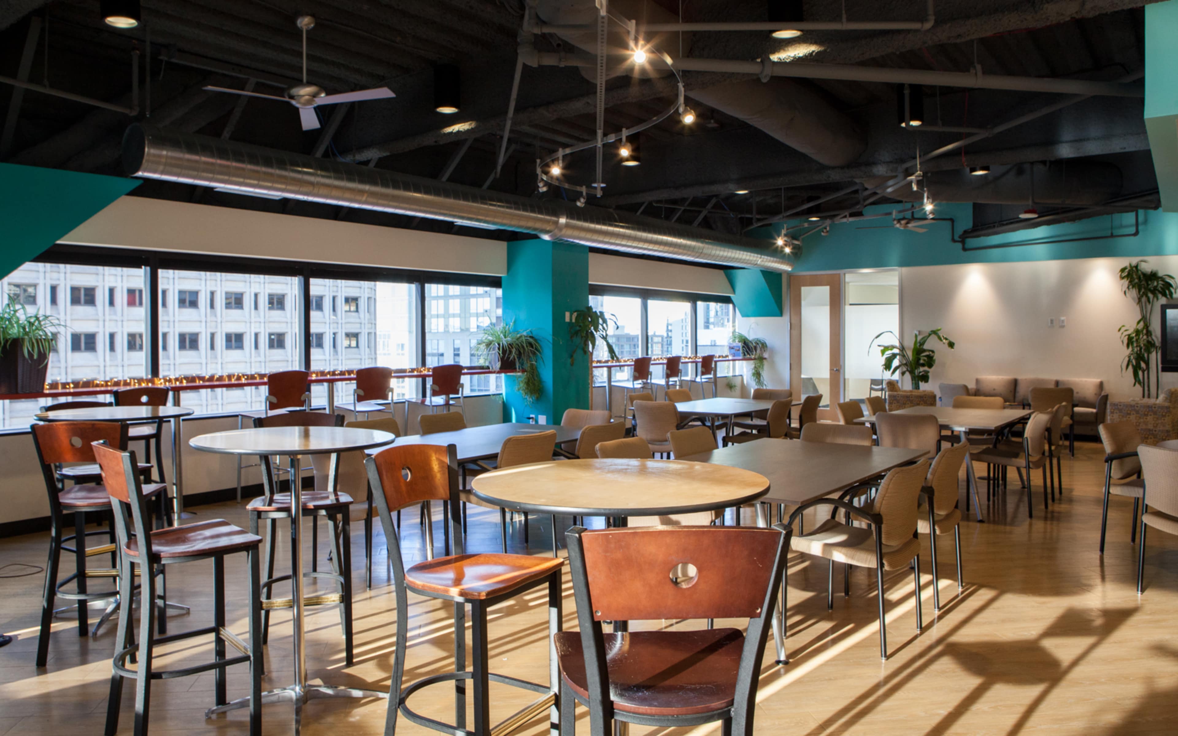 A spacious indoor dining area with multiple round and rectangular tables surrounded by chairs, large windows letting in natural light, and greenery in pots along the walls.