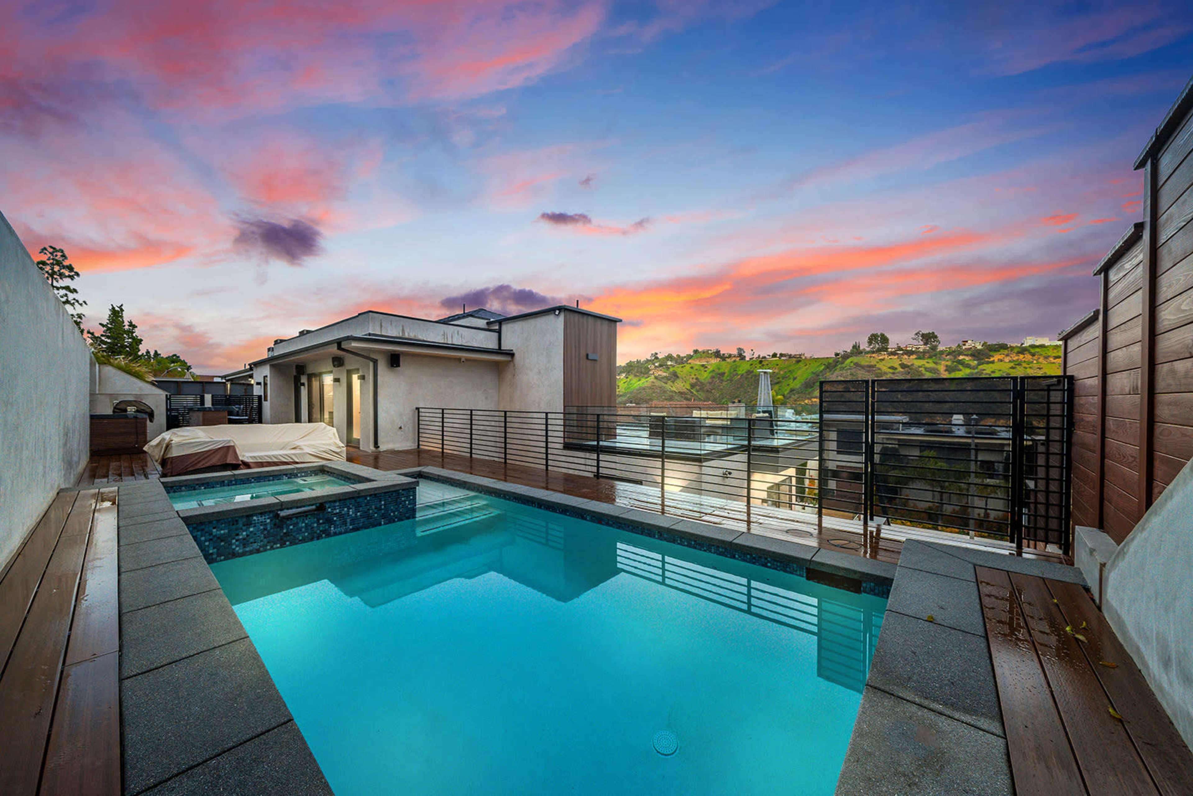 A modern rooftop pool surrounded by wooden decking and a view of hills under a colorful sunset sky.