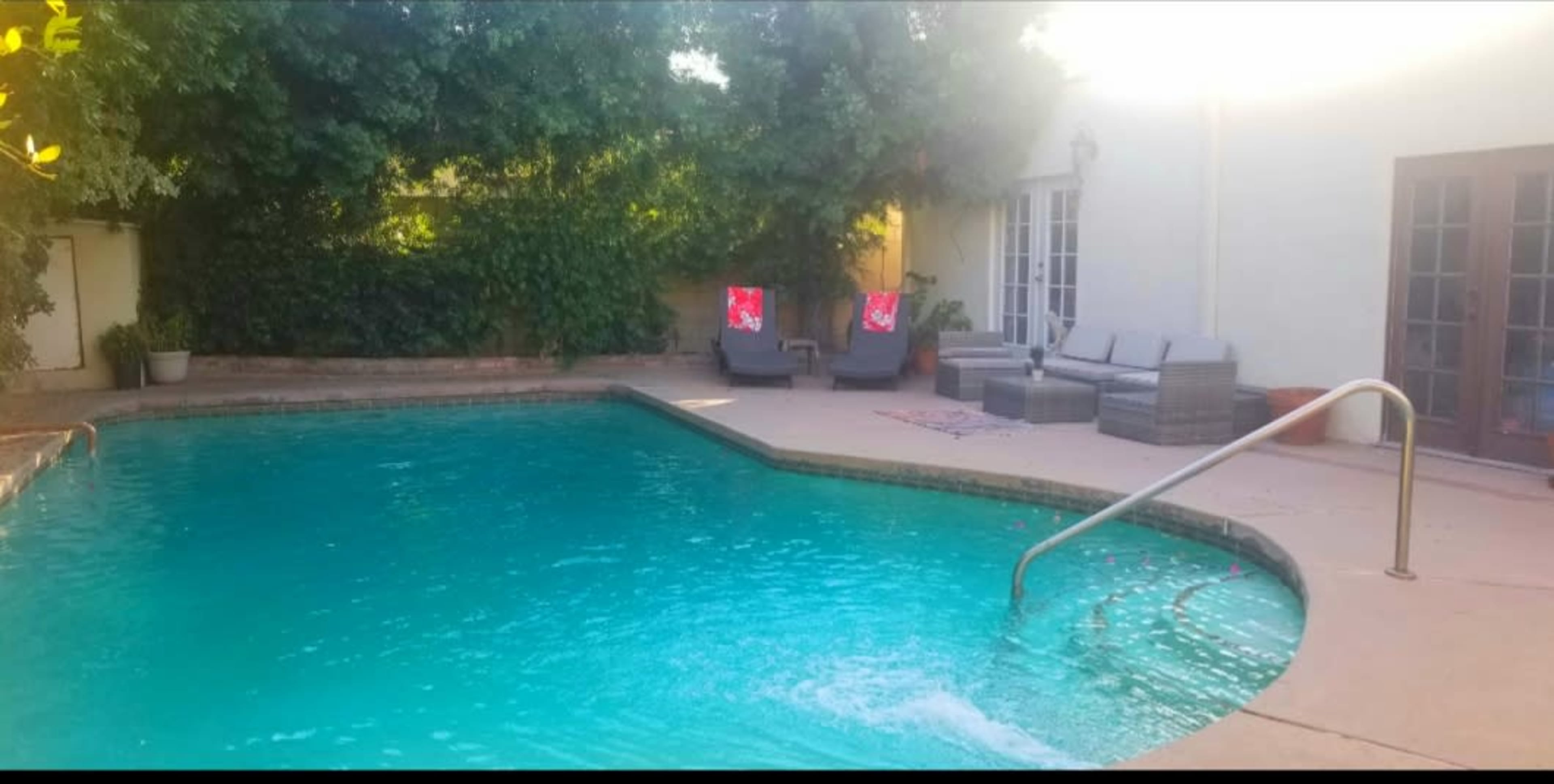 A swimming pool surrounded by green foliage, with lounge chairs and a small table nearby.