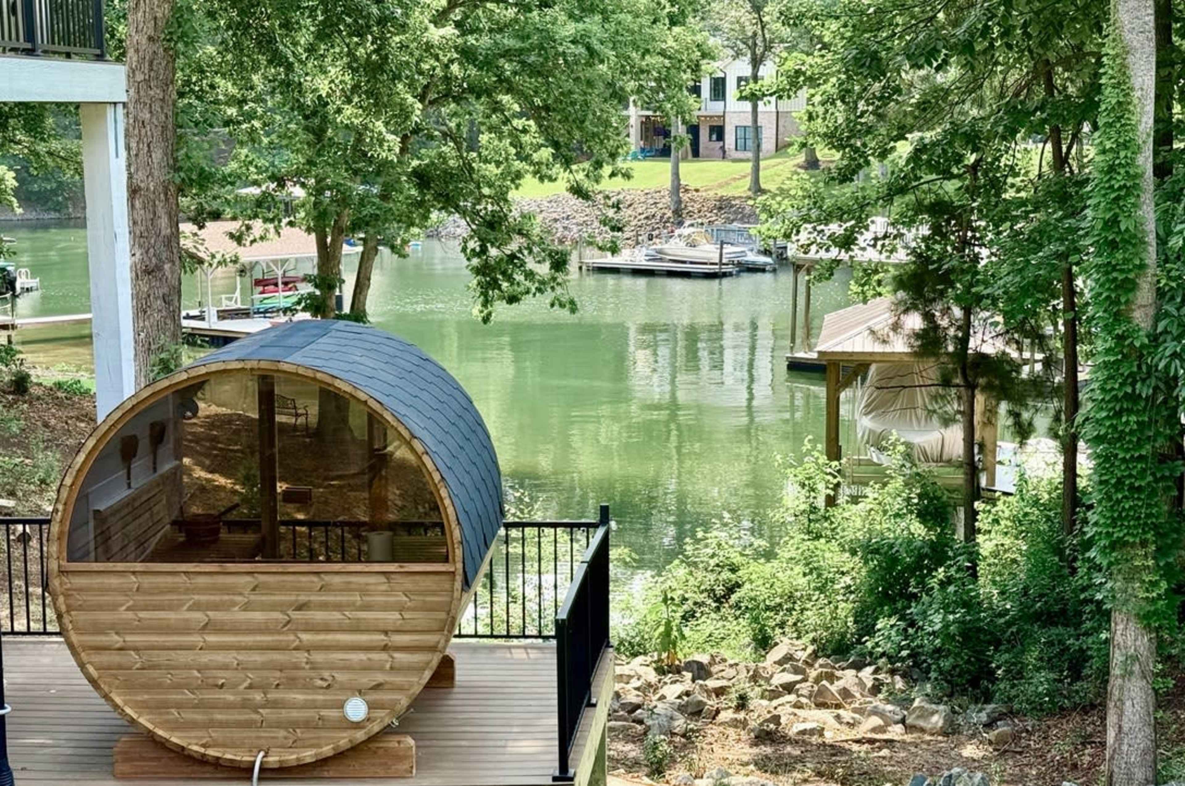 A wooden barrel-shaped sauna is positioned on a deck overlooking a calm lake surrounded by greenery and waterfront cottages.