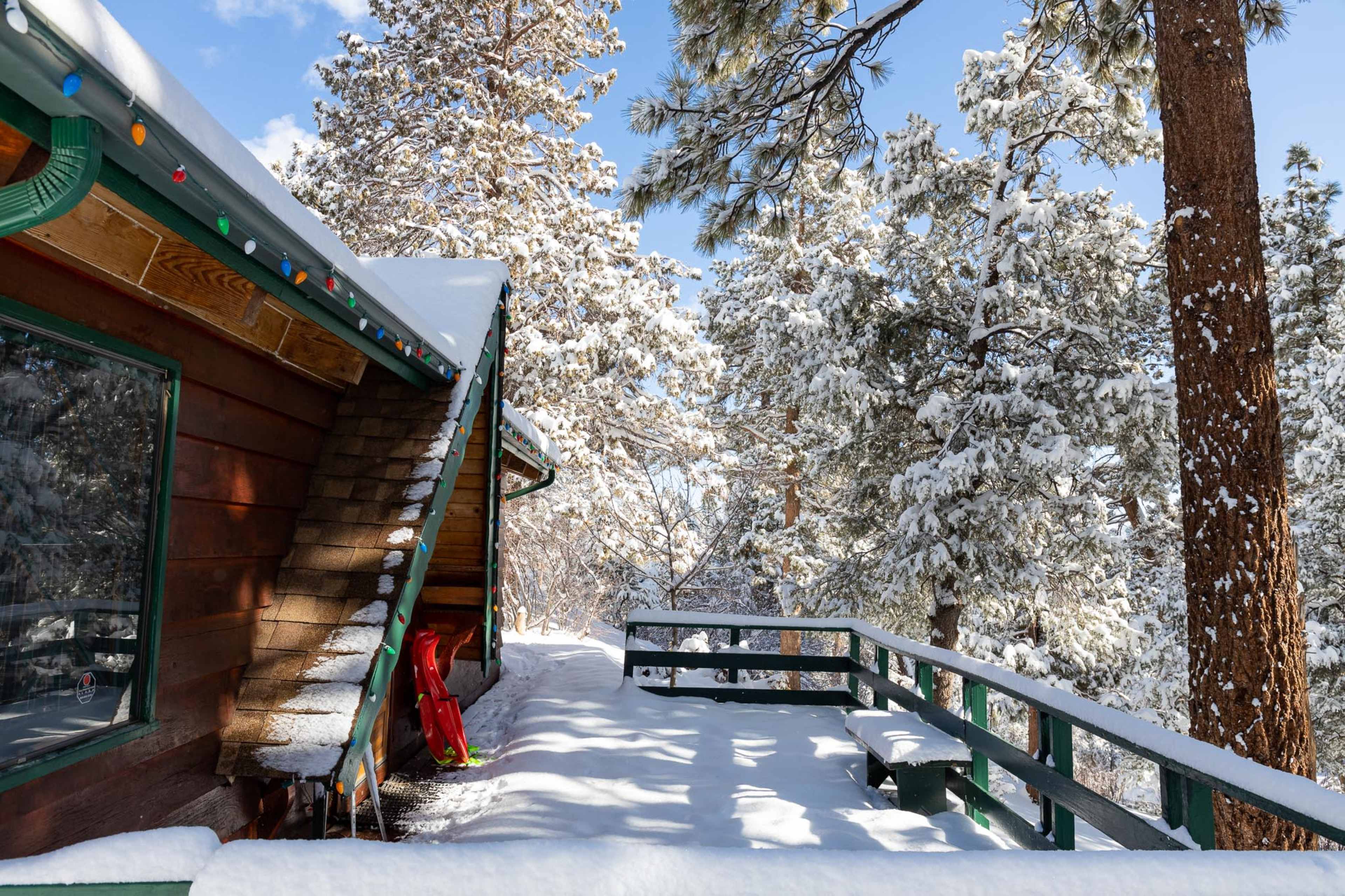 A wooden cabin partially covered in snow is nestled among tall evergreen trees under a clear blue sky.