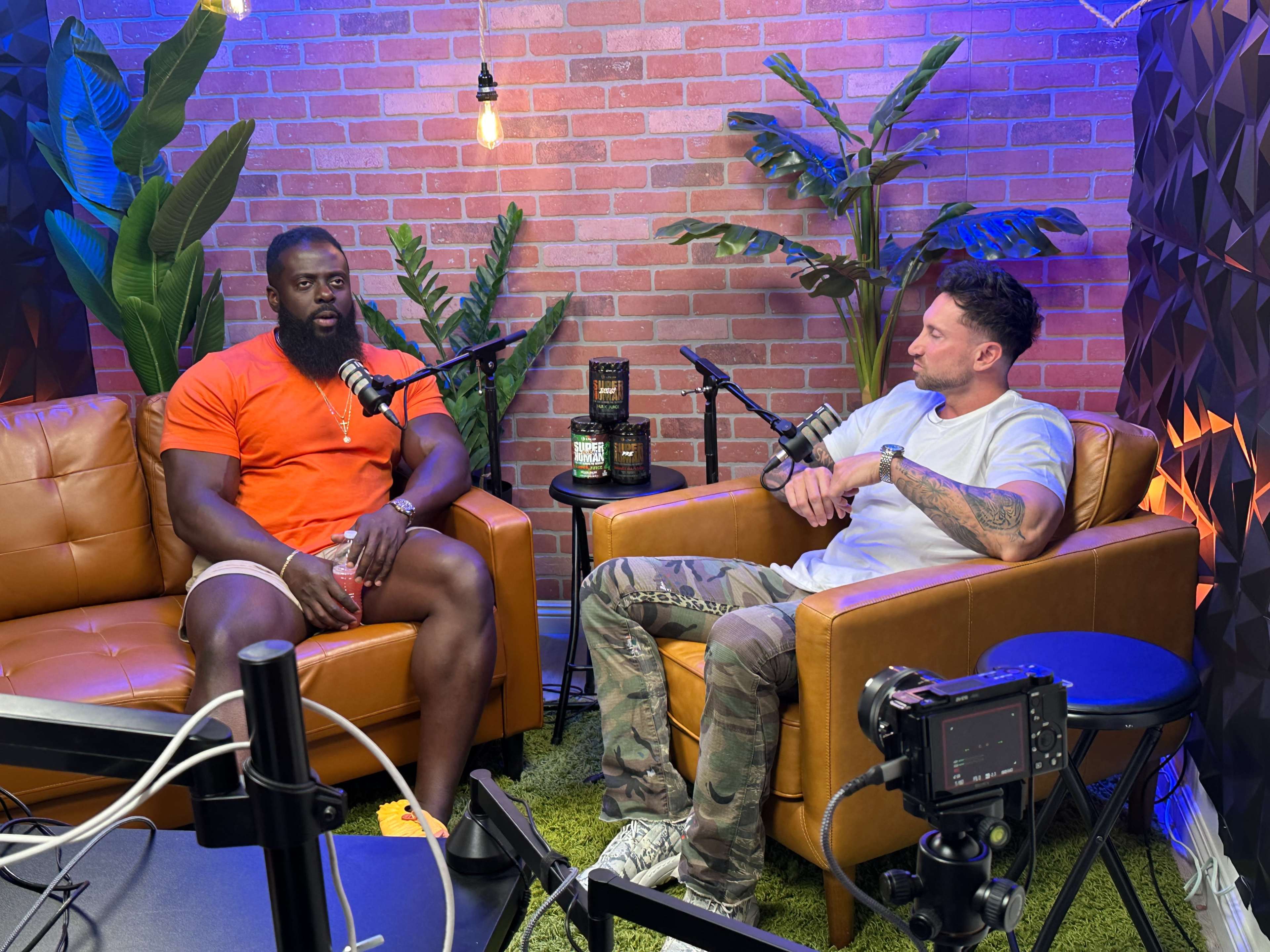 Two men sit in armchairs in a podcast studio, discussing into microphones, with plants and decorative lighting in the background.