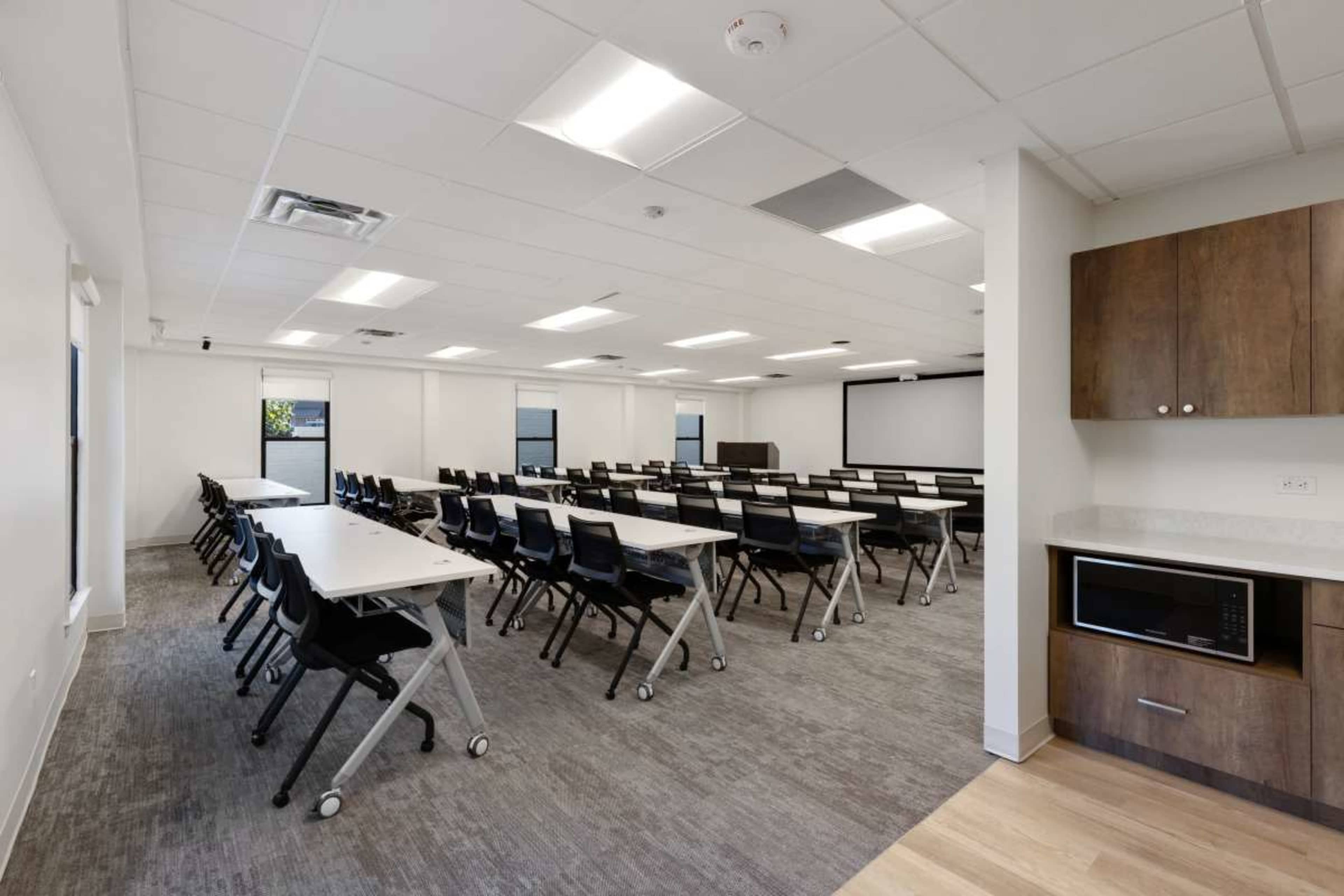 The image shows a modern empty classroom with rows of tables and chairs, featuring a kitchen area with a microwave in the corner.