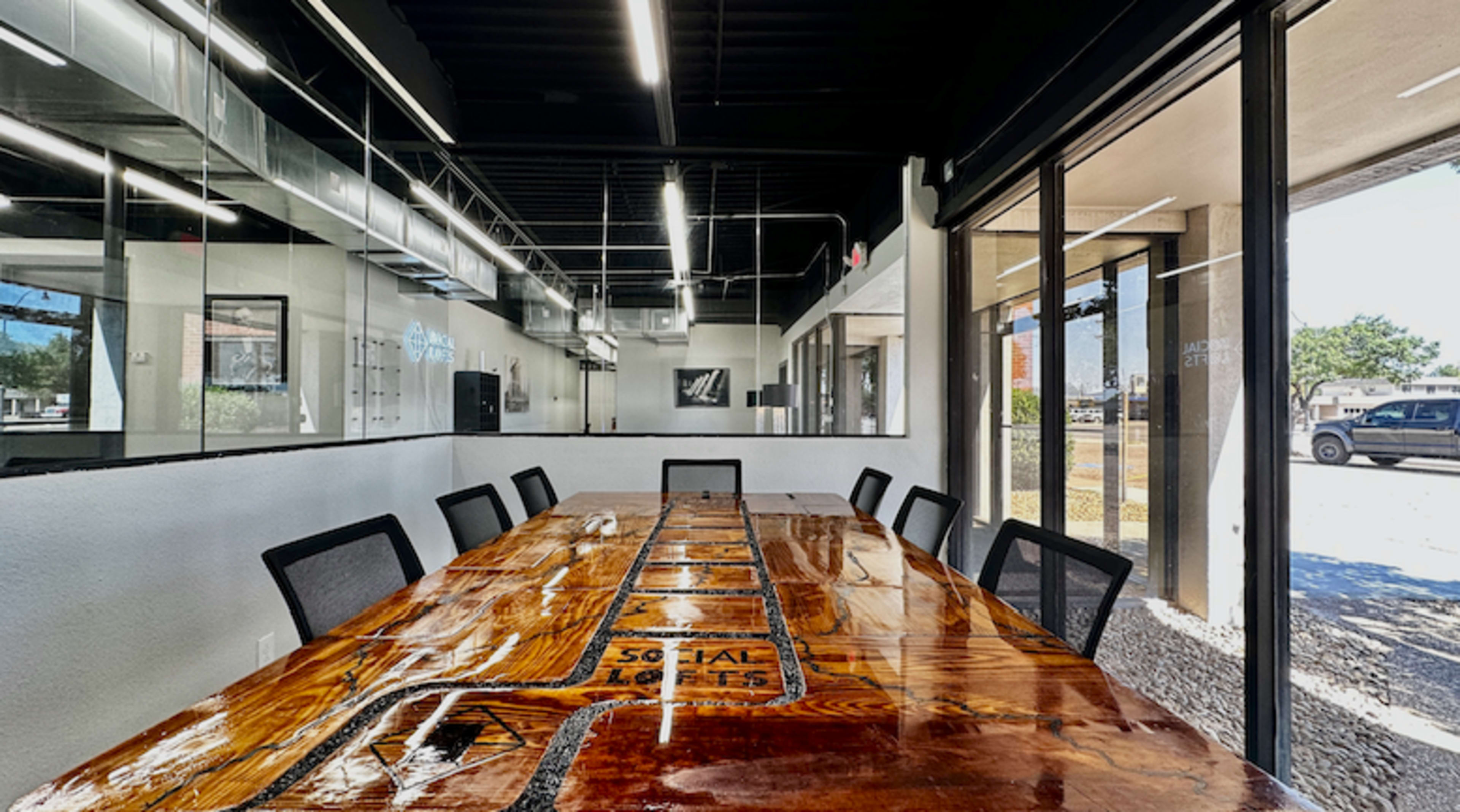 The image shows a modern conference room featuring a large wooden table with a glossy finish and a row of black chairs arranged around it, with large glass walls and a view of the exterior.