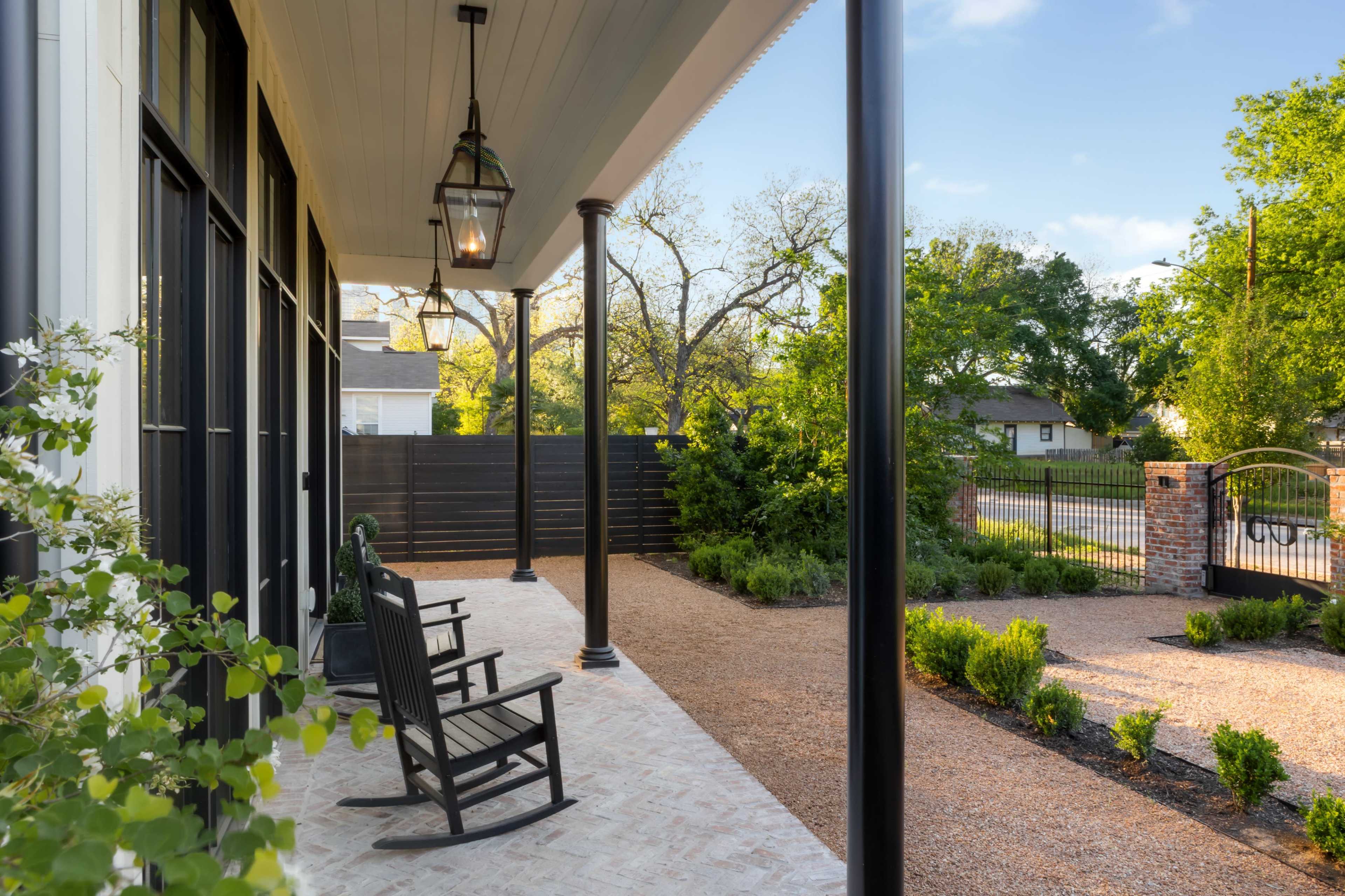 The image shows a covered porch with rocking chairs overlooking a gravel pathway and landscaped greenery.