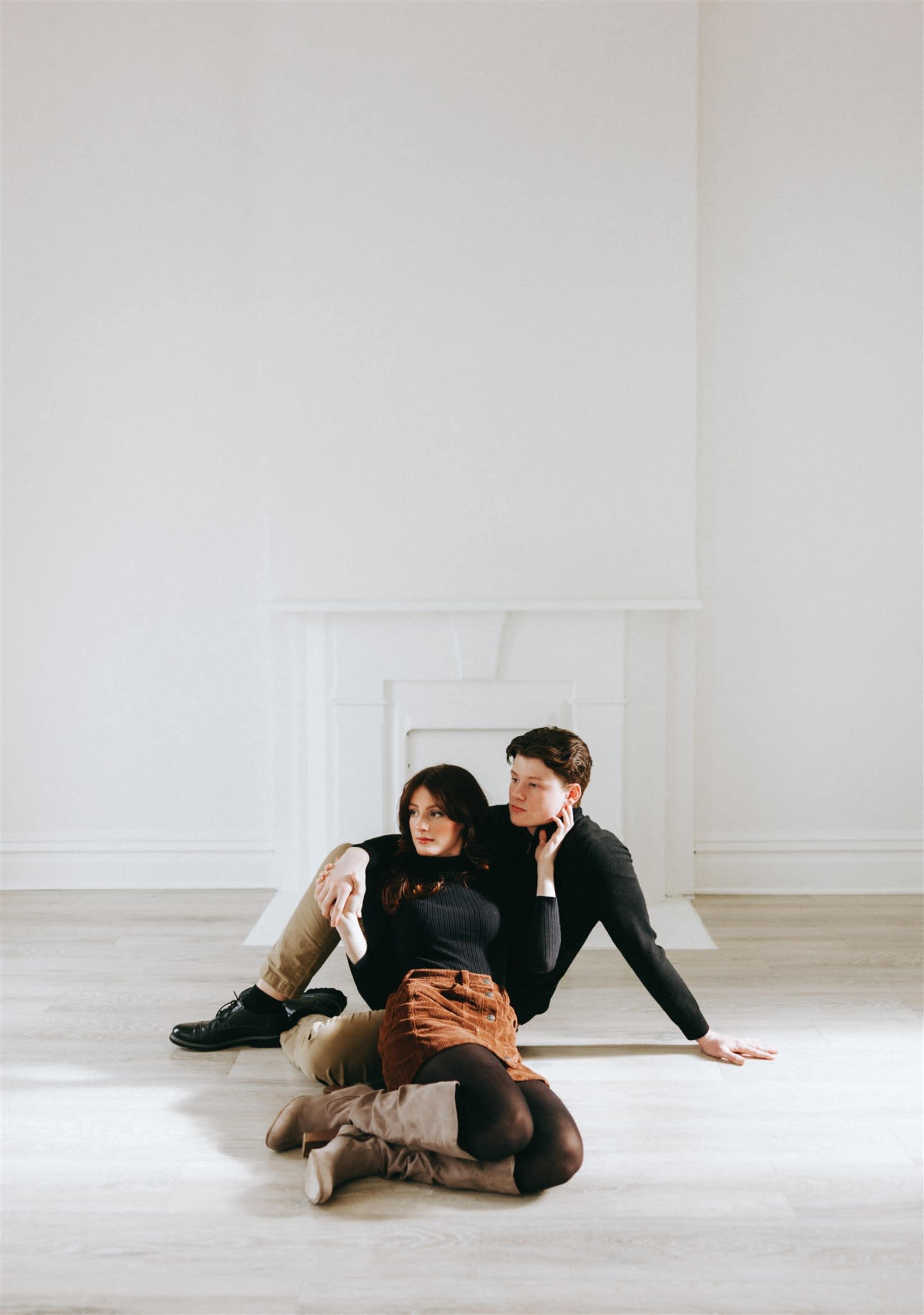 Two individuals sit on the floor of a brightly lit, minimalistic room with a white fireplace in the background.