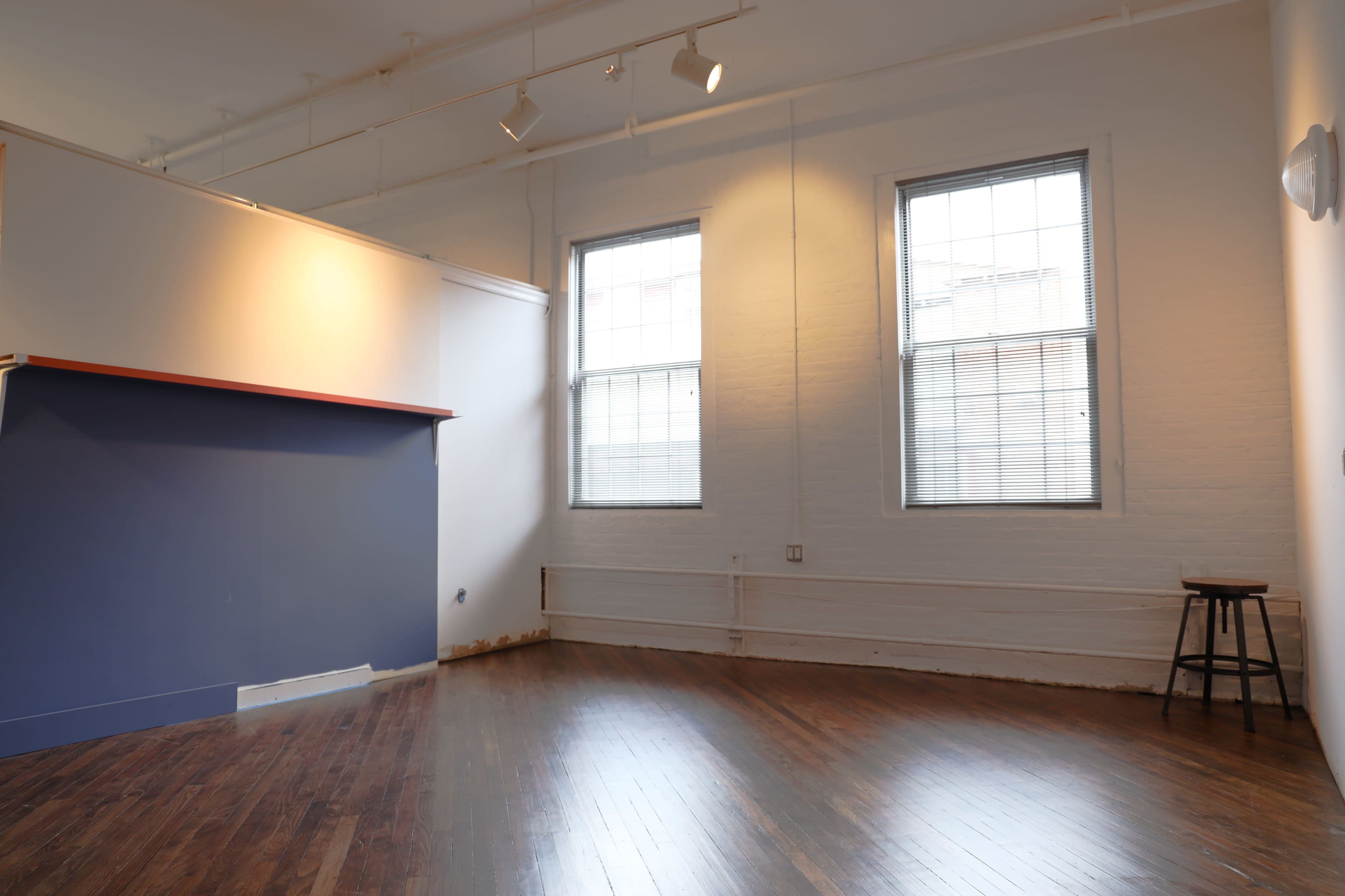 An empty room with hardwood floors, two windows, a blue wall section, and a wooden stool in the corner.