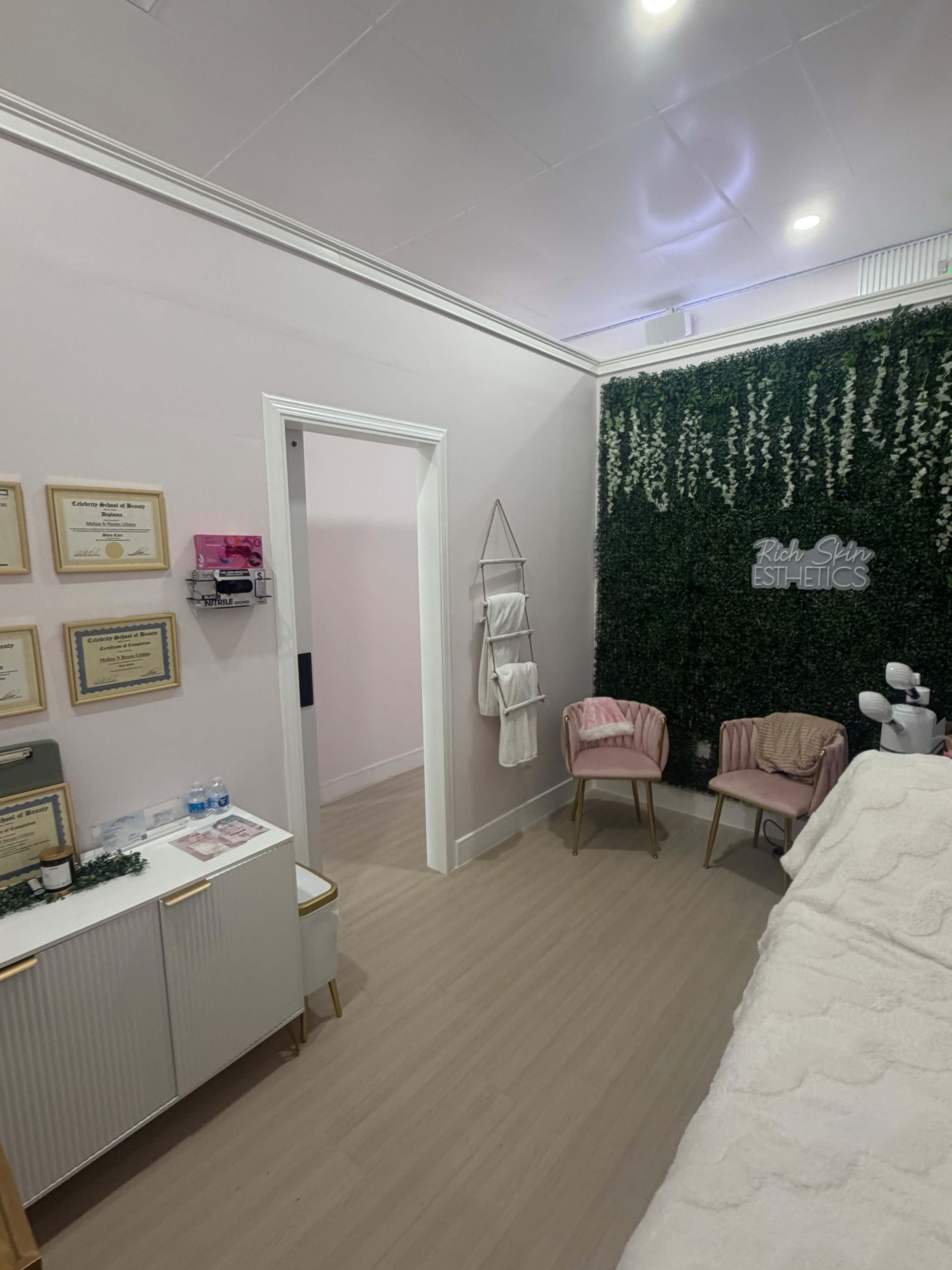 The image shows a small, elegantly designed esthetics room with pink walls, a green wall covered in artificial plants, and two pink chairs beside a cabinet displaying framed certificates.